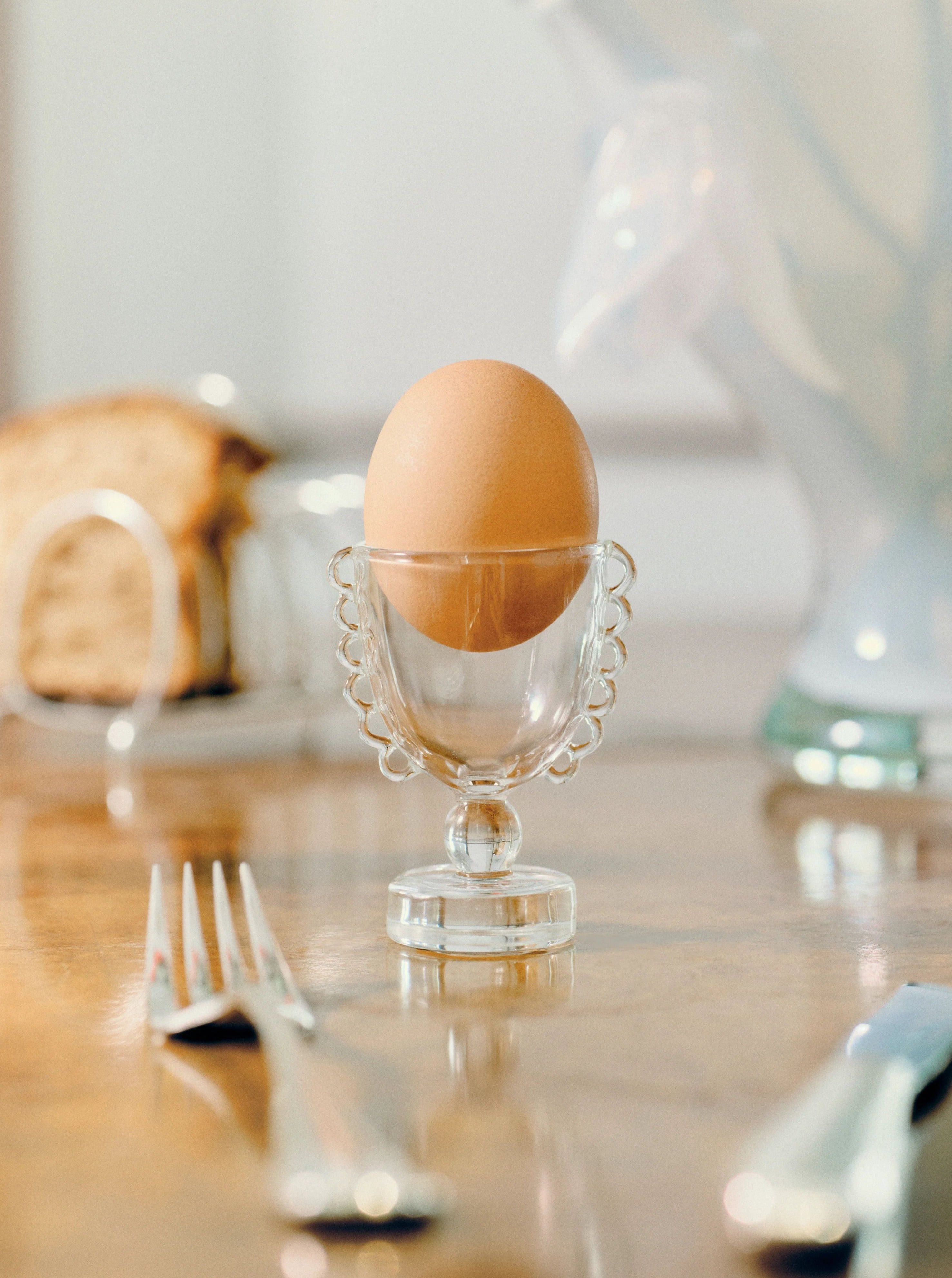 A brown egg sits elegantly in a delicate, transparent Coco Egg Cup Set by Sophie Lou Jacobsen with decorative handles, resembling a miniature shot glass. The egg is the focal point, with blurred slices of bread on a holder and cutlery in the background, creating a refined and minimalist breakfast setting.