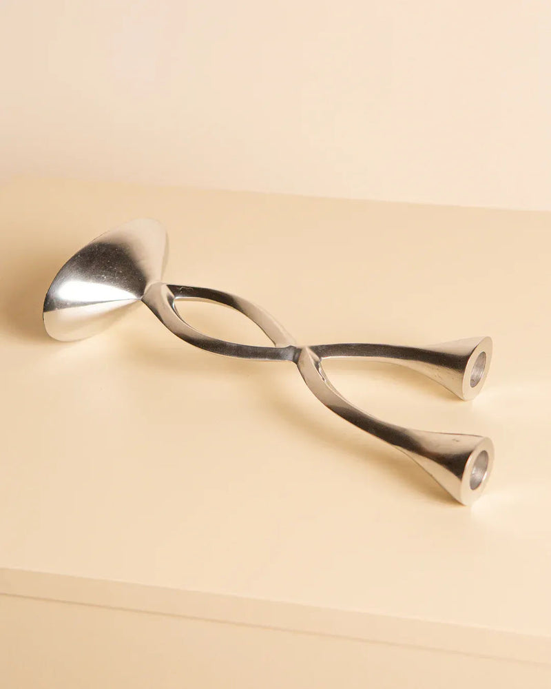 The "Torsadé" 80's Candleholder by Treaptyque features a modern, abstract aluminum design resembling interwoven, rounded arms on one side and a flat, oval shape on the other. It sits elegantly on a light-colored surface against a minimalist backdrop.
