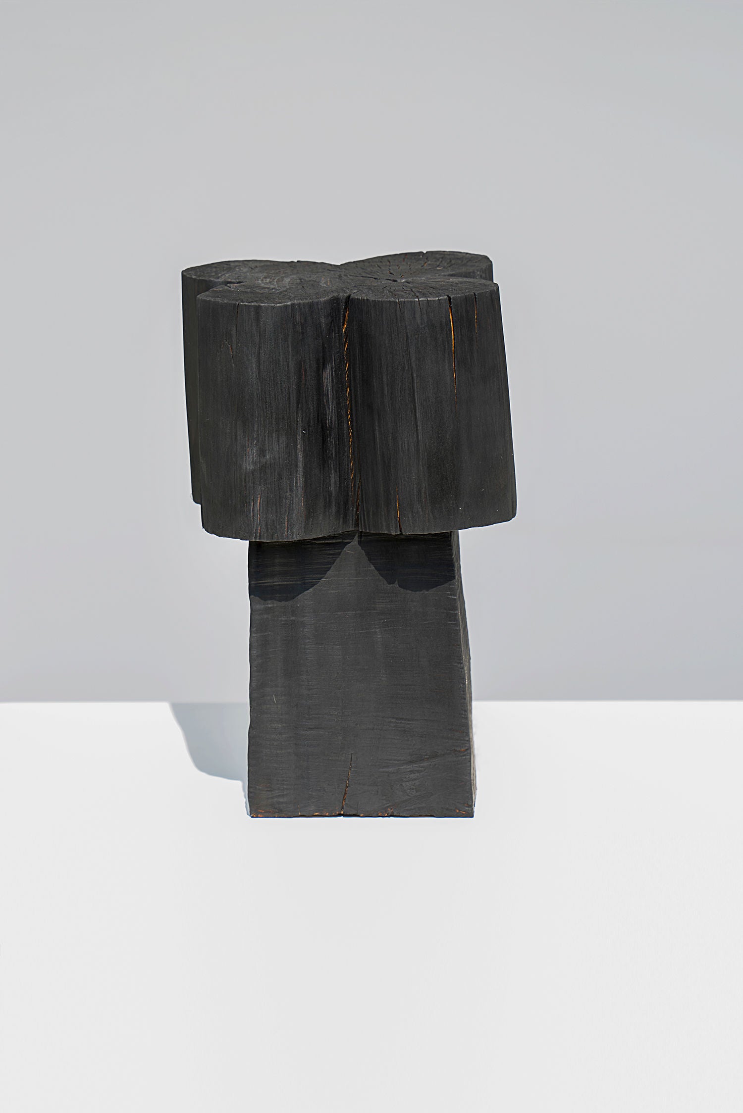 The Burnt Black Coffee Tables/Stools by Touch With Eyes is a minimalist, handmade wood sculpture with a charred finish, featuring a round top and rectangular base. It stands on a white surface against a light gray background, reminiscent of modern black tables.