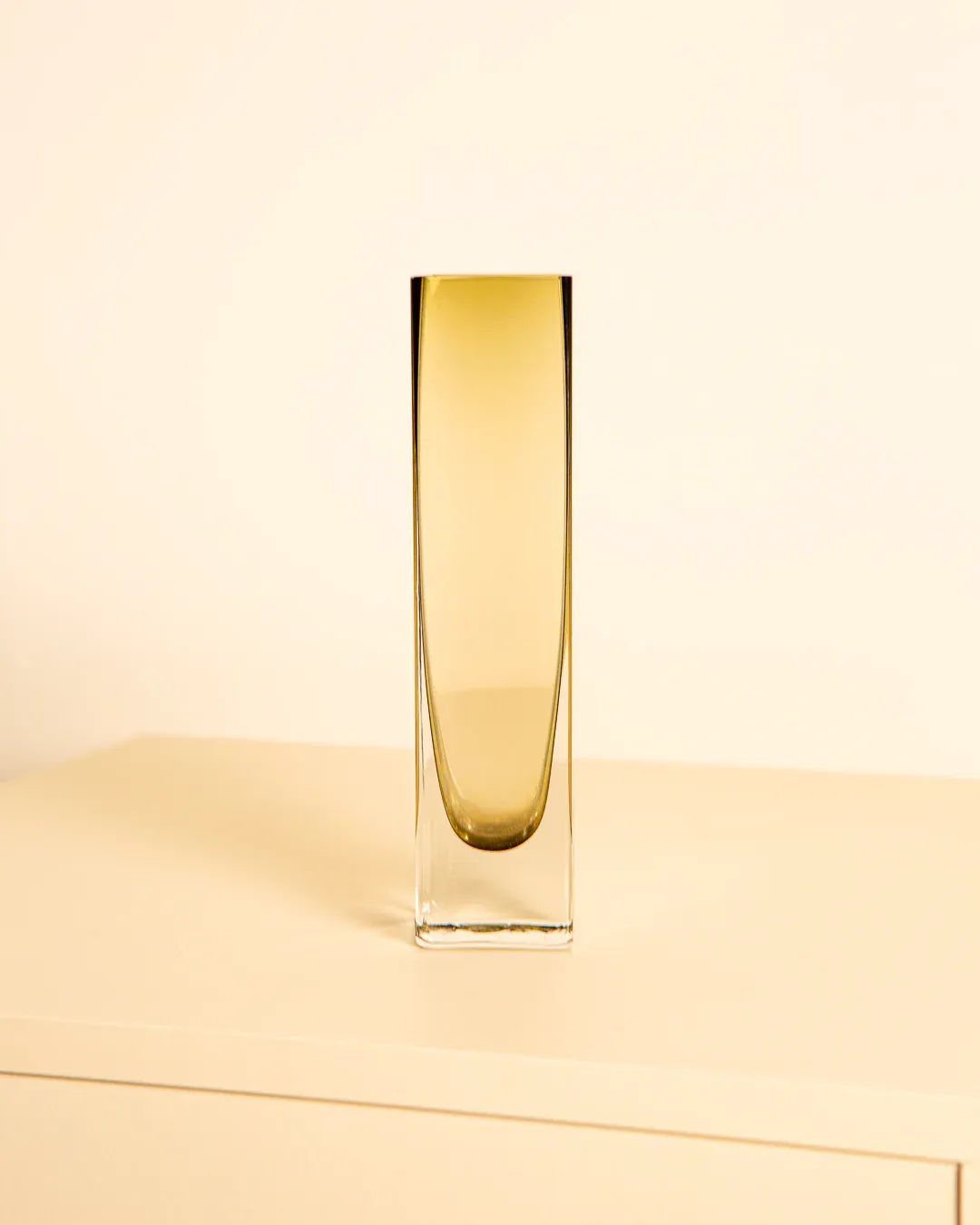 The Treaptyque 80's Murano Glass Amber Vase, tall and slender with a gradient from clear to amber, exudes vintage elegance on a light surface against a pale background.