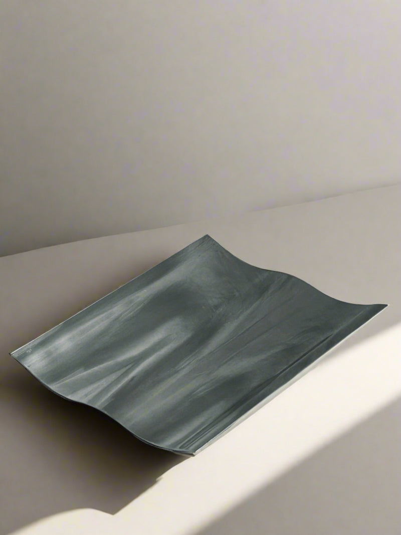 A Wave Tray by Royal Copenhagen, crafted by Flock Studio, is placed on a plain surface. The sheet's folds and curves create a modern twist on design, while its angle captures soft light that casts shadows and highlights its unique texture.