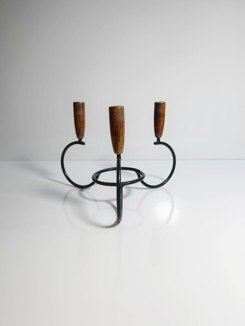 A minimalist black metal three-arm Danish Candleholder 1960s by Dodo Vintage, featuring teak ends, is placed on a smooth, reflective white surface against a light grey background. The design showcases clean lines and a modern aesthetic.