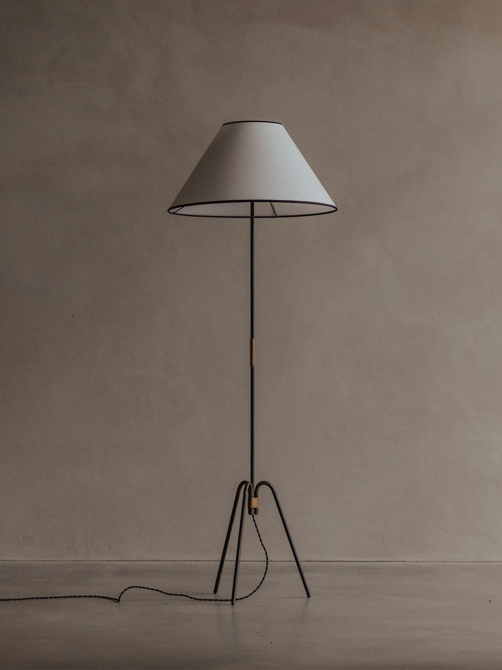 The Bicci de' Medici Manner Of Royere & Adnet Wrought Iron Tripod Lamp, with a sleek black metal stand and white conical shade, stands on a gray floor against a beige wall, showcasing subtle mid-20th-century design elements.