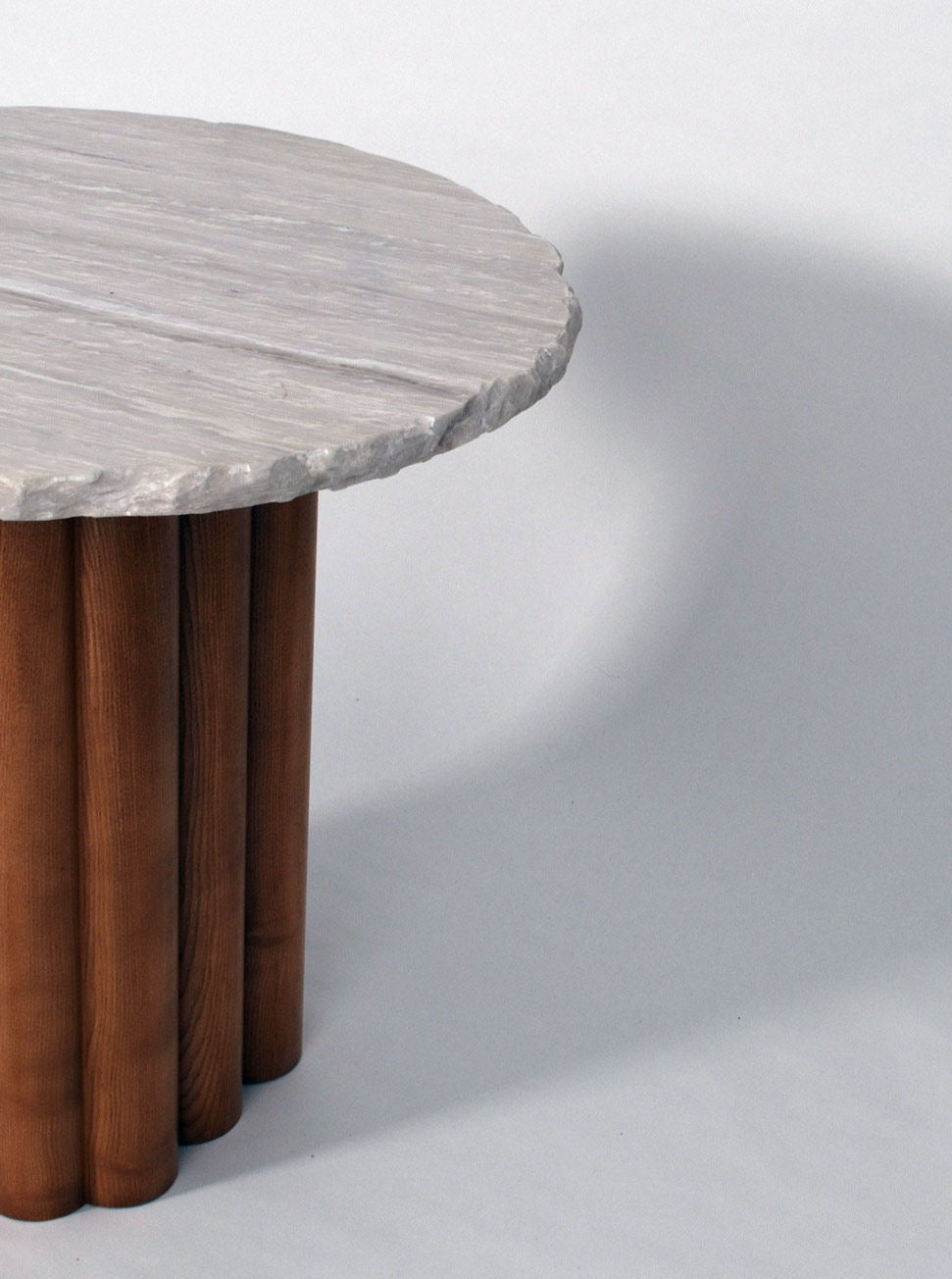 A small, round table with a textured white marble top and a base made of cylindrical oak legs is positioned on a neutral, light-colored background. Shadows from the table are cast on the surface below. This is the "Carrasqueira" table, Barracuda edition by Barracuda Interiors.