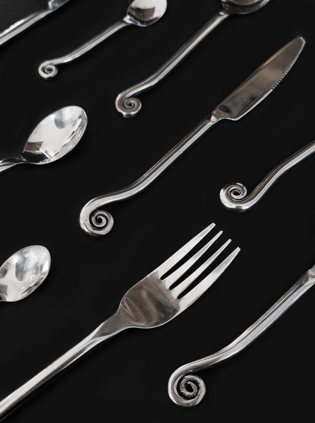 The Set of 5 Swirling Cutlery by Les Objoies is displayed on a black background, featuring silver spoons, forks, and knives with unique curled spiral designs at the handles.