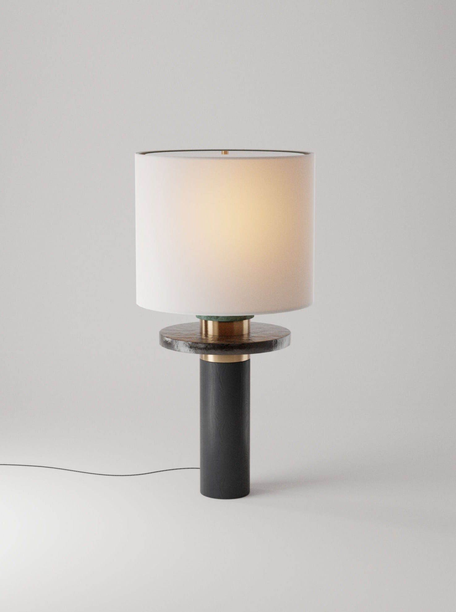 A modern Ines table lamp by Marine Breynaert with a cylindrical solid brass base and a wide, circular white lampshade, illuminated, set against a plain light grey background.