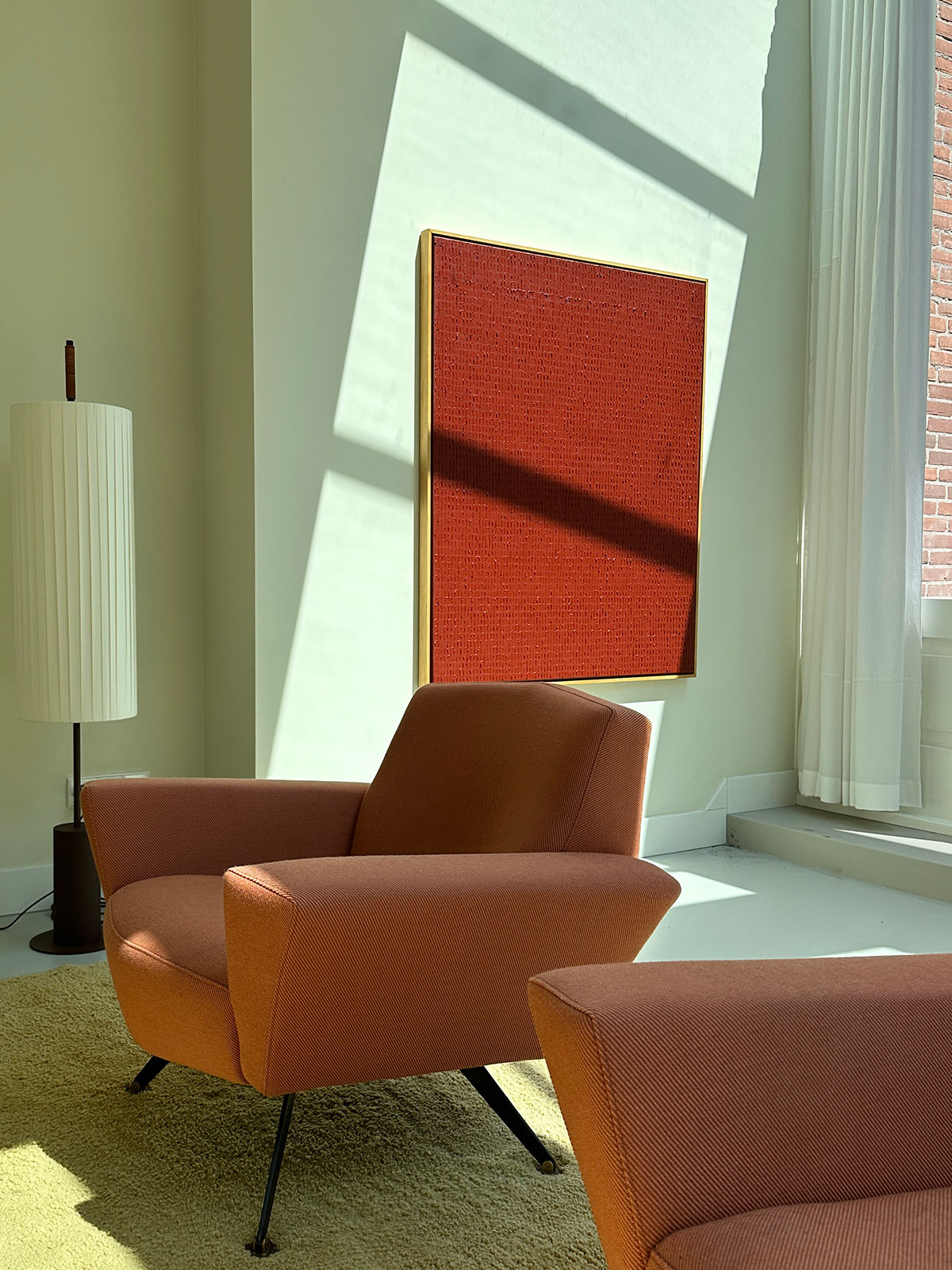 A sunlit room with modern furniture, featuring two orange armchairs on a light beige rug. Behind the chairs, "Indian Red - Oil on canvas" by Roi Elmaliah hangs on the wall. A tall, cylindrical white lamp stands to the left of the painting. Sunlight streams through large windows.