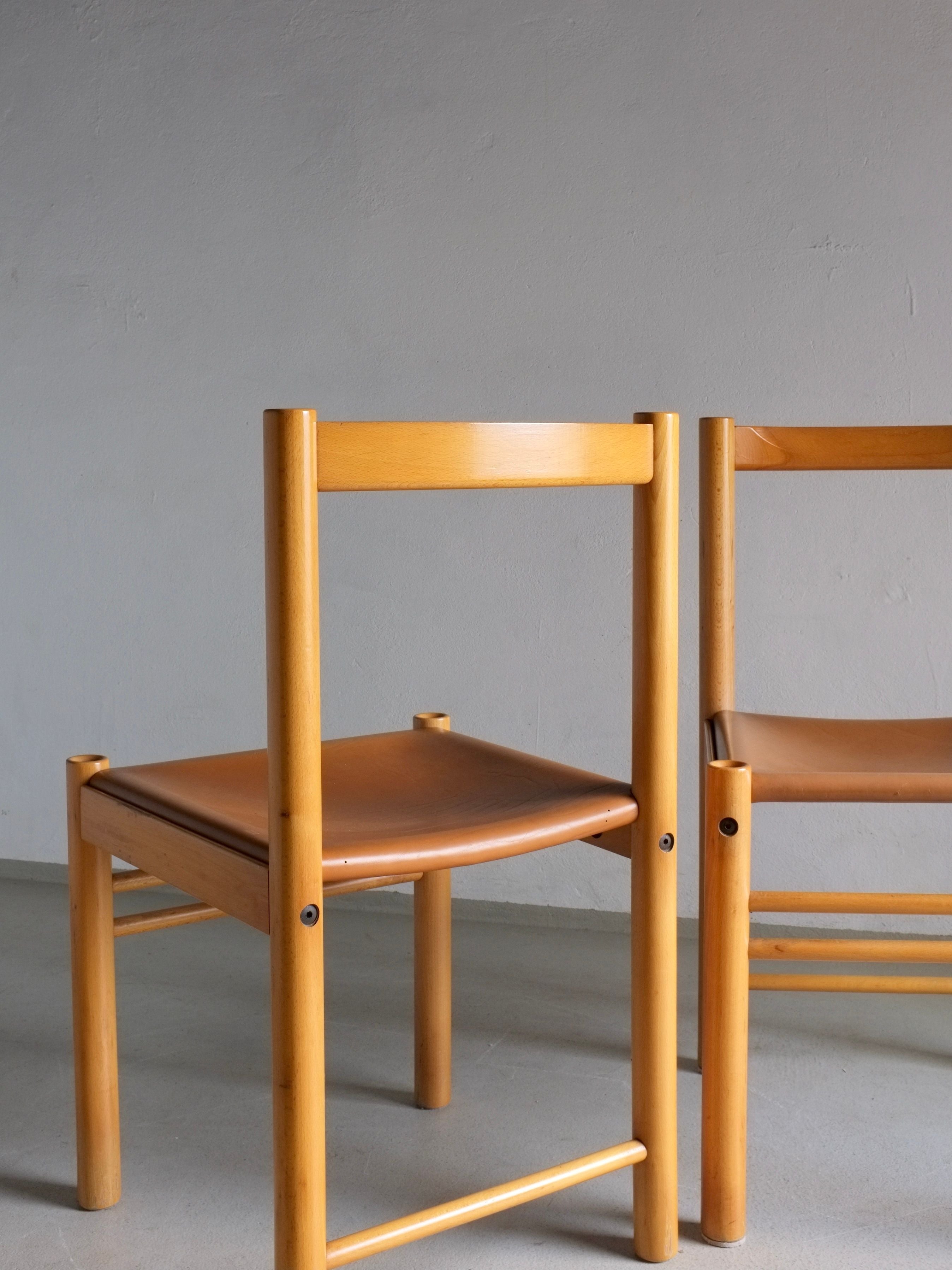 Six Veter Vintage Ibisco dining chairs from the 1960s showcase a simple, modern design with straight beech wood legs and rectangular backrests. The seats are upholstered in cognac leather and set against a plain gray wall, perfectly capturing a minimalist, timeless aesthetic.