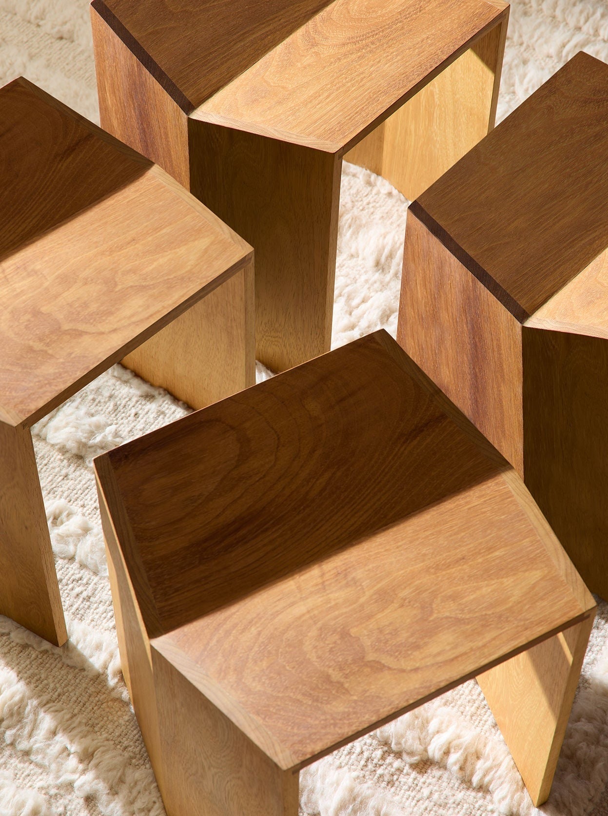 Several wooden hexagonal Lemon Athens Stools in Iroko arranged artistically on a cream-colored shaggy rug, showing varying solid Iroko wood grains and shades under soft lighting.