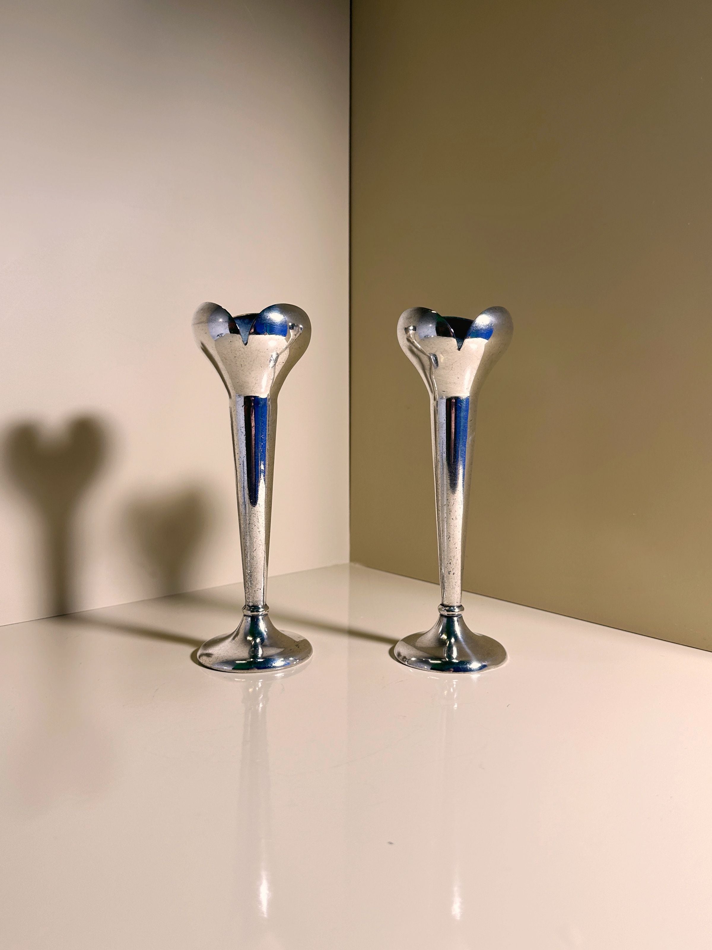 Two silver, heart-shaped candlesticks by Gio Ponti for Calderoni from the 1930s stand side by side on a beige surface. Their reflections form heart-shaped shadows, enhancing the elegant and romantic ambiance. Available at Dodo Vintage.