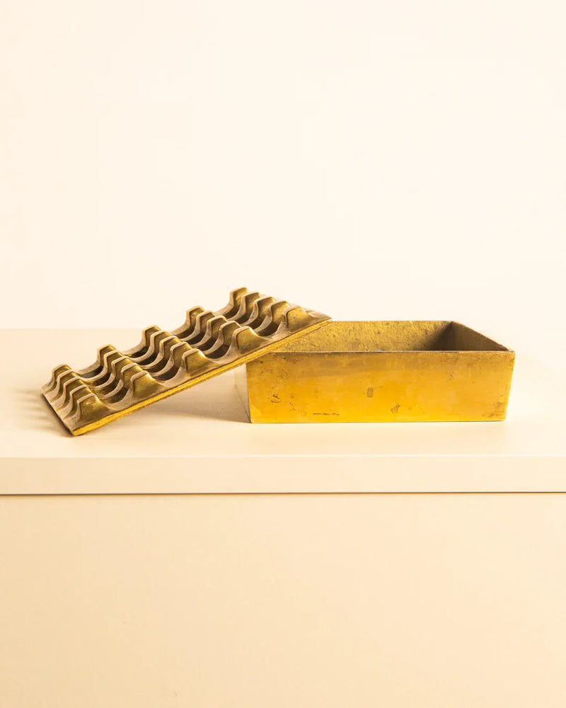 A vintage yellow metal ashtray, crafted from high-quality brass by Holger Bäckström for Treaptyque, boasts an open rectangular base and a removable lid with wavy ridges. This elegant piece rests gracefully on a white surface against a light background.