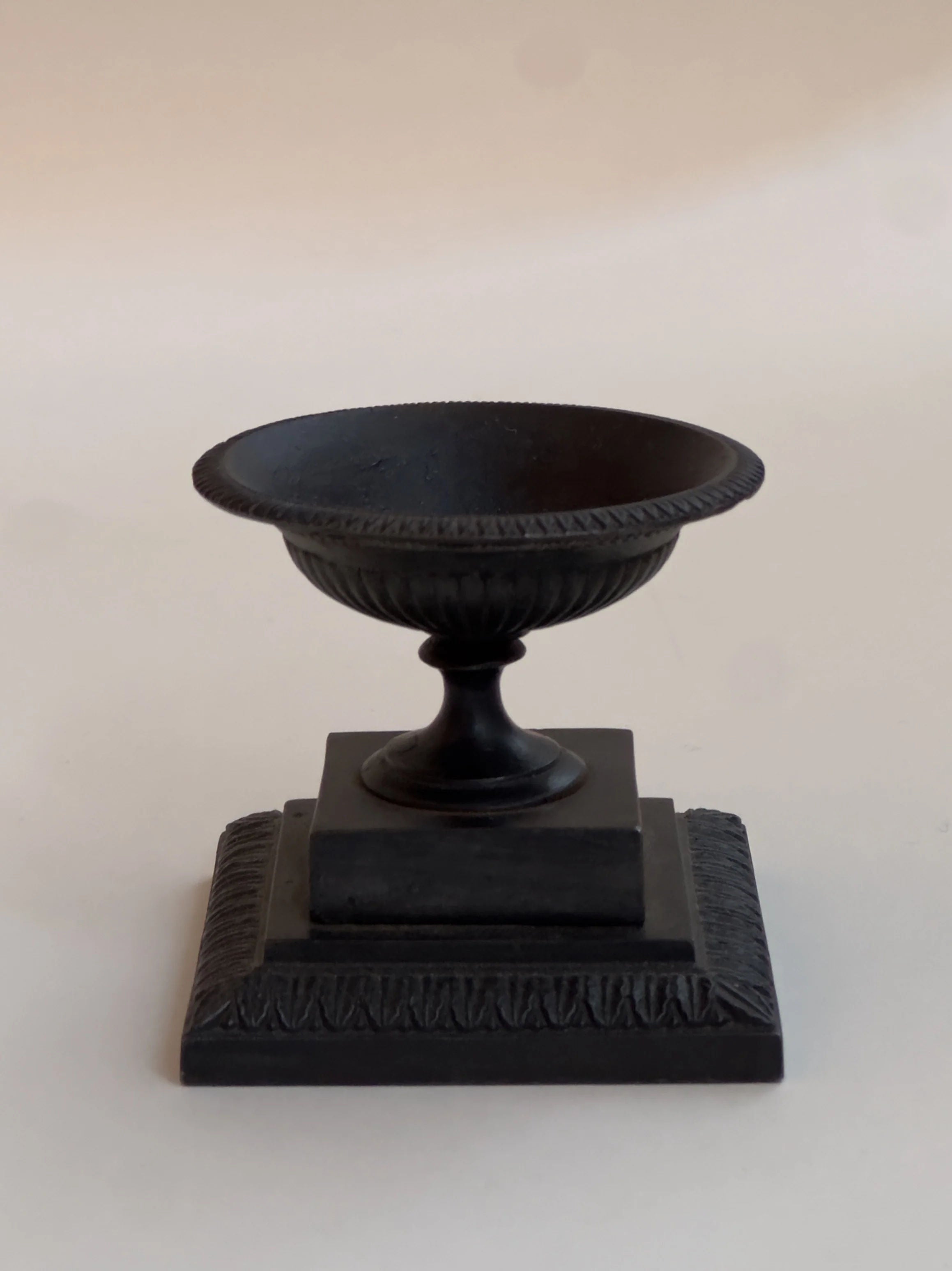 The Small Cast Iron Urn by Galerie Storm features a black, chalice-shaped design reminiscent of an antique tazza urn. It elegantly sits on a pedestal with a textured square base, showcasing ornate detailing that exudes classic 1800s charm against a light background.