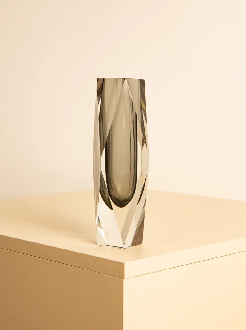A sleek, transparent crystal vase with faceted edges stands on a minimalist beige surface, set against a plain off-white background. This Black “Diamond” vase by Flavio Poli for Mandruzzato 70s, reminiscent of Italian glass art brought to you by Treaptyque, features elongated vertical facets that create a modern and elegant aesthetic.
