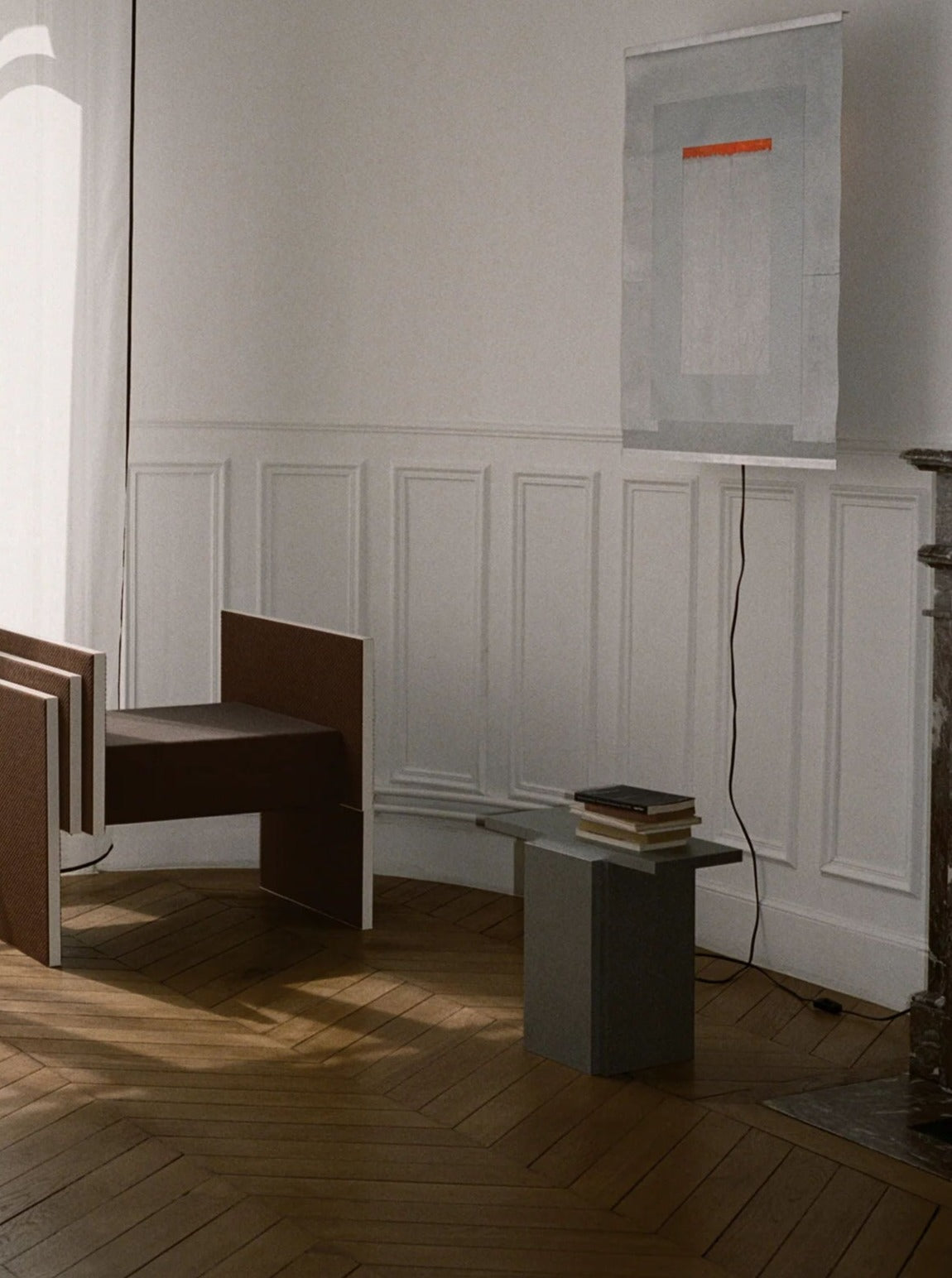 A minimalist room features a modern chair paired with TEGET's "Mara T" Side Table in Silver Noise, elegantly holding books on a wooden floor. The wall is adorned with decorative paneling and a contemporary art piece in orange and textured gray hues. A fireplace is partially visible on the right as natural light filters in.