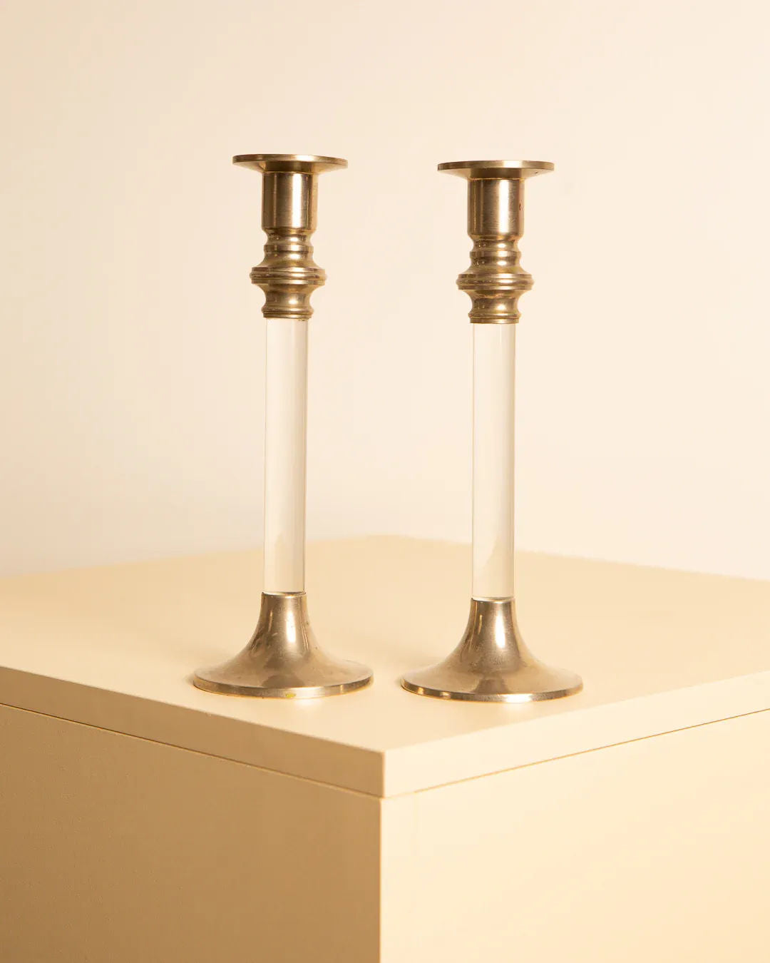 Two elegant Treaptyque Pair of 80's silver and lucite candleholders, reminiscent of retro candleholders, are placed side by side on a beige surface. The design features a silver base and top with a clear lucite center, highlighting a minimalist and sophisticated aesthetic.