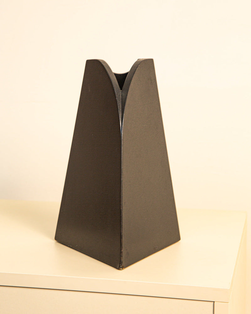 The Treaptyque Italian “Triangle” Vase 1980s, featuring a modern matte black finish and split curved top, is displayed on a light surface with a plain off-white background.