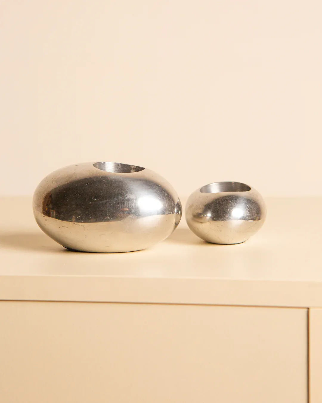 A pair of "Galet" candleholders from Treaptyque, with their shiny, oval-shaped silver designs, rest on a light-colored surface against a beige background. These aluminum pieces embody minimalist design with organic shapes, adding elegance to any space.