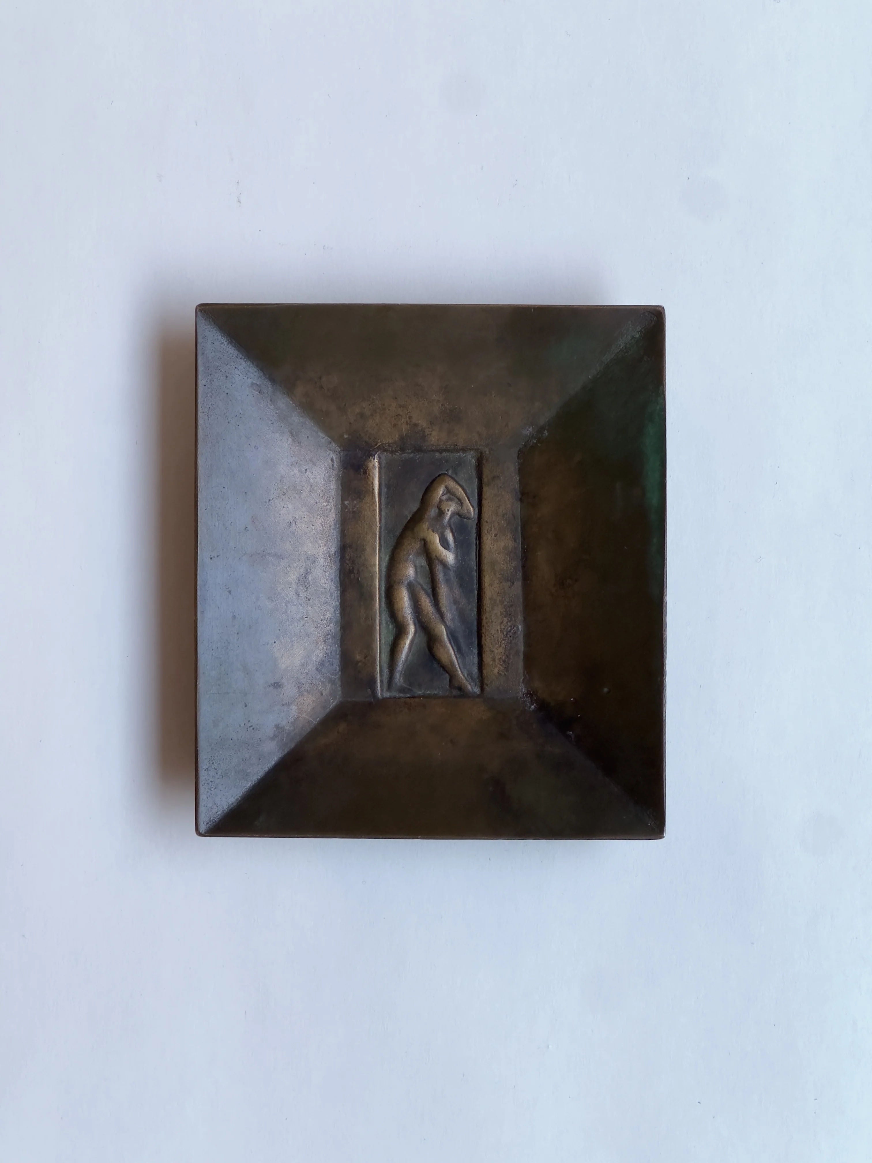 Galerie Storm's Bronze dish, GAB, by Jacob Ängman displays a dynamic center figure with sloped edges that lead the eye inward, casting intriguing shadows on its patina-rich surface against a plain white backdrop.