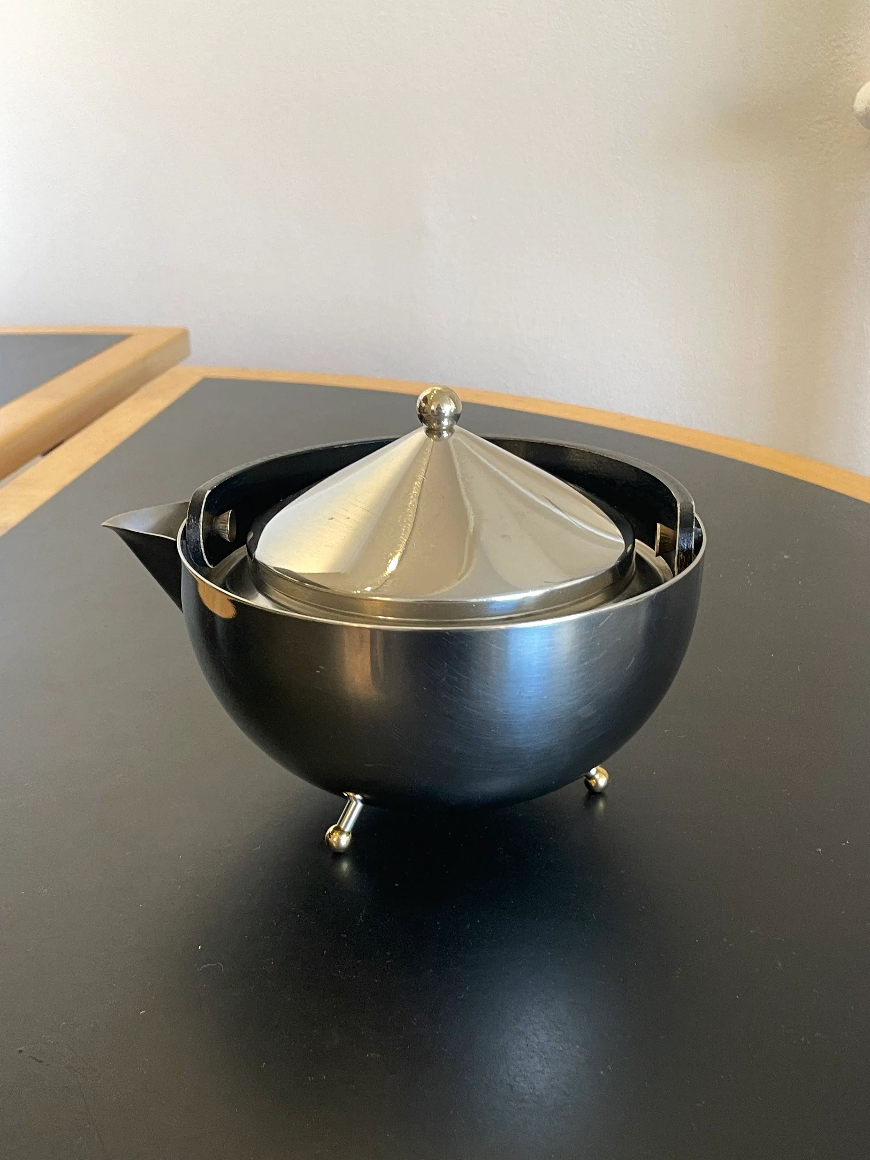 A silver, dome-shaped Collect Cph Teaball Teapot with a polished lid and an elegant spout is positioned on a dark countertop. The teapot stands on three small, spherical feet and reflects light, adding a touch of modernity with its sleek stainless steel design.