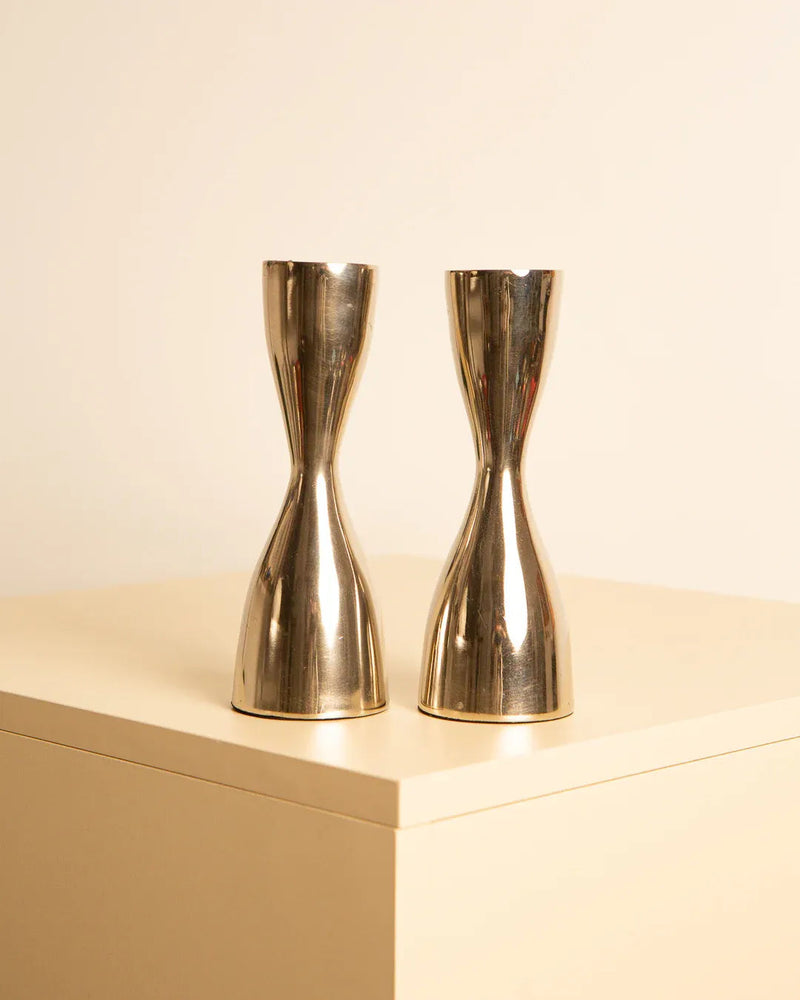 Two gold Hourglass candleholders in Italian metal 80's by Treaptyque, reminiscent of 1980s Italy, rest on a beige surface against a plain background. They have a reflective, polished finish and appear identical in size and shape.