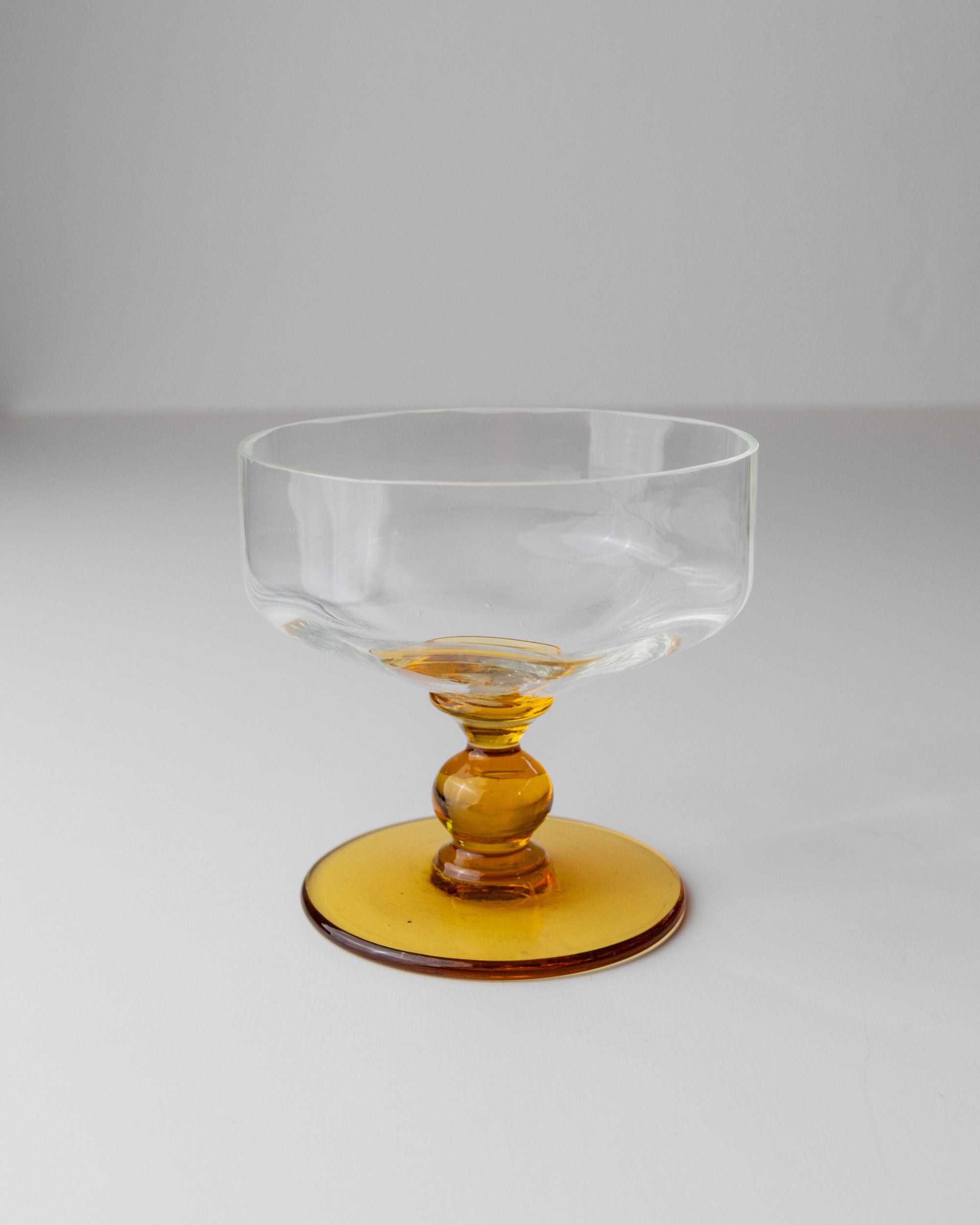 A set of six Bottega Jacobs Murano glass bowls from the 70s, featuring clear glass with wide rims and short stems, sit on round amber bases reminiscent of vintage style, displayed on a plain white surface.