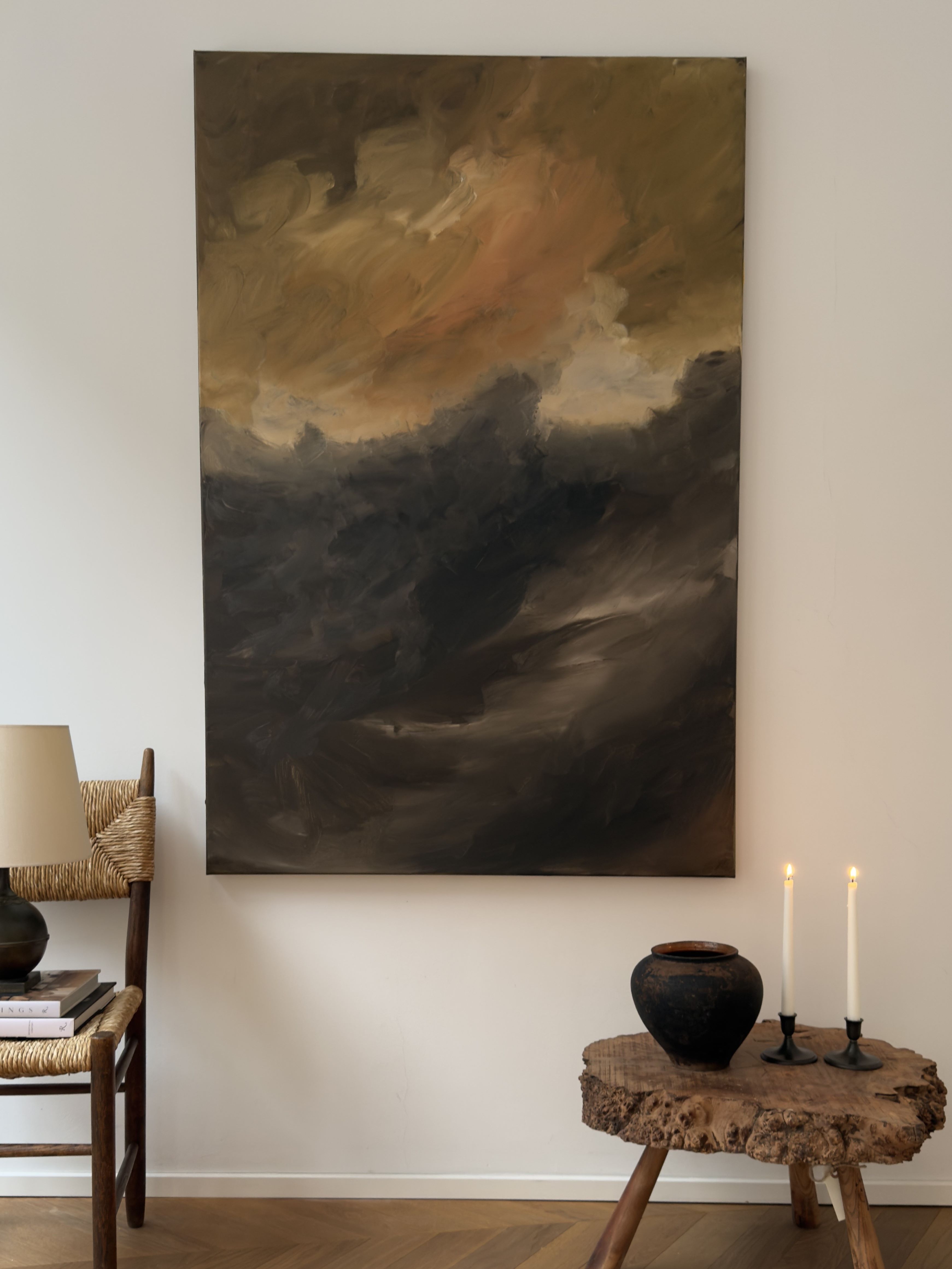 The "Whispering Peaks" painting by Mattias Storm, featuring large abstract designs with dark, swirling colors, is mounted on a wall above a rustic wooden table. The table holds a black vase and two lit candles. Nearby on the wooden floor rests a woven chair accompanied by a lamp and stacked books, telling tales of stillness and art.