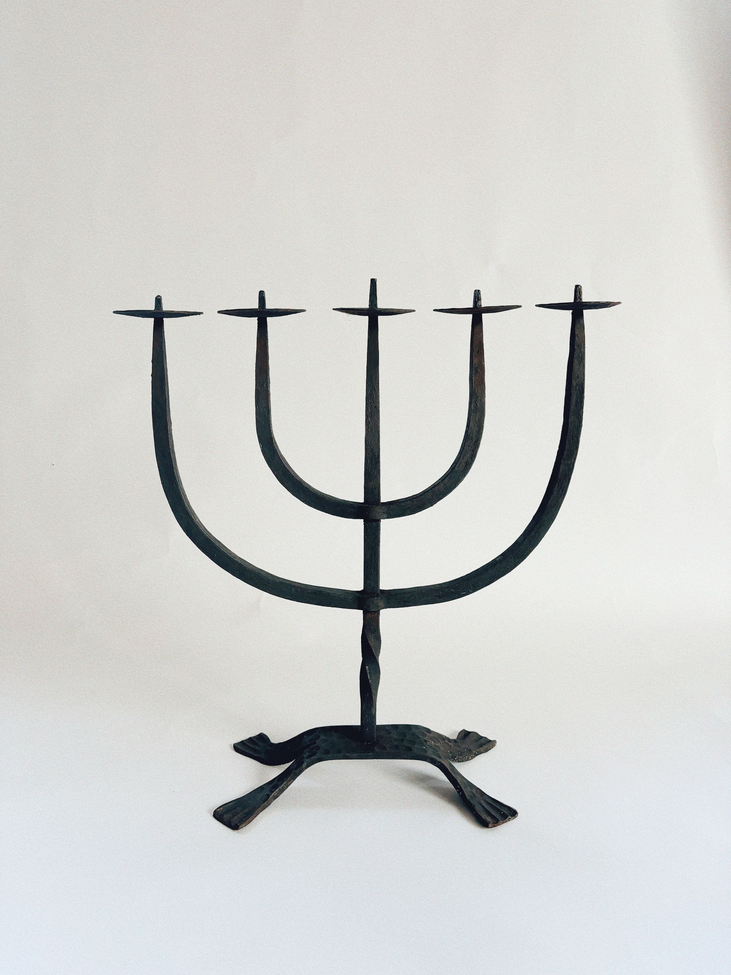 The Huerto Hand Forged Candleholder features a minimalist design with five branches, each holding a candle atop an elegant iron structure. Artisan crafted, this candelabra boasts a twisted wrought iron motif for its base, providing stable support against a light background.