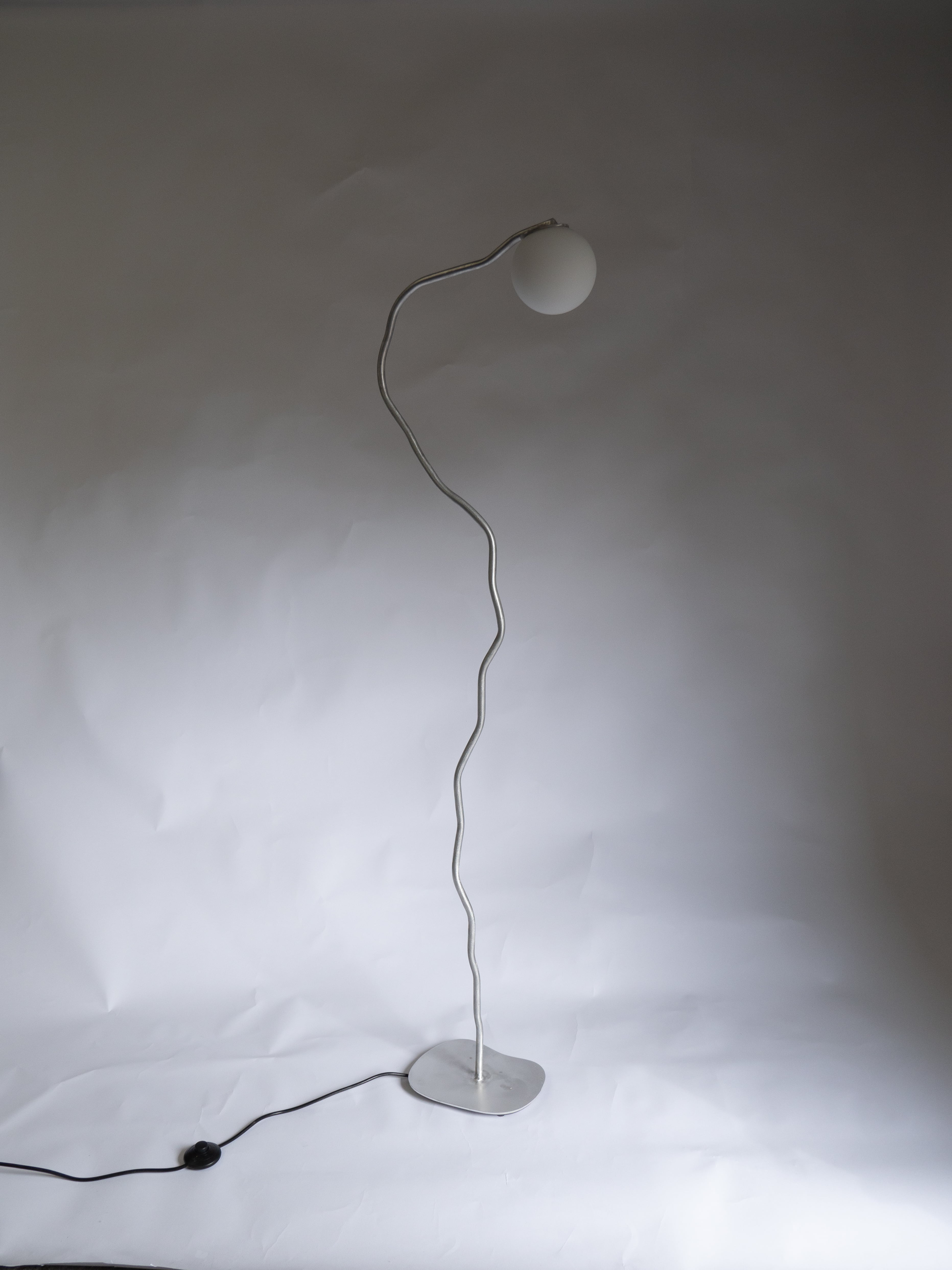 A modern, handcrafted floor lamp with a uniquely curved, wavy silver stand and a spherical white lampshade at the top. It stands on a stable, flat base and has a black power cord extending from the base. The Six Dots Design Pea Head Floor Lamp sits against a plain, light-colored surface, adding natural ambience to any room.