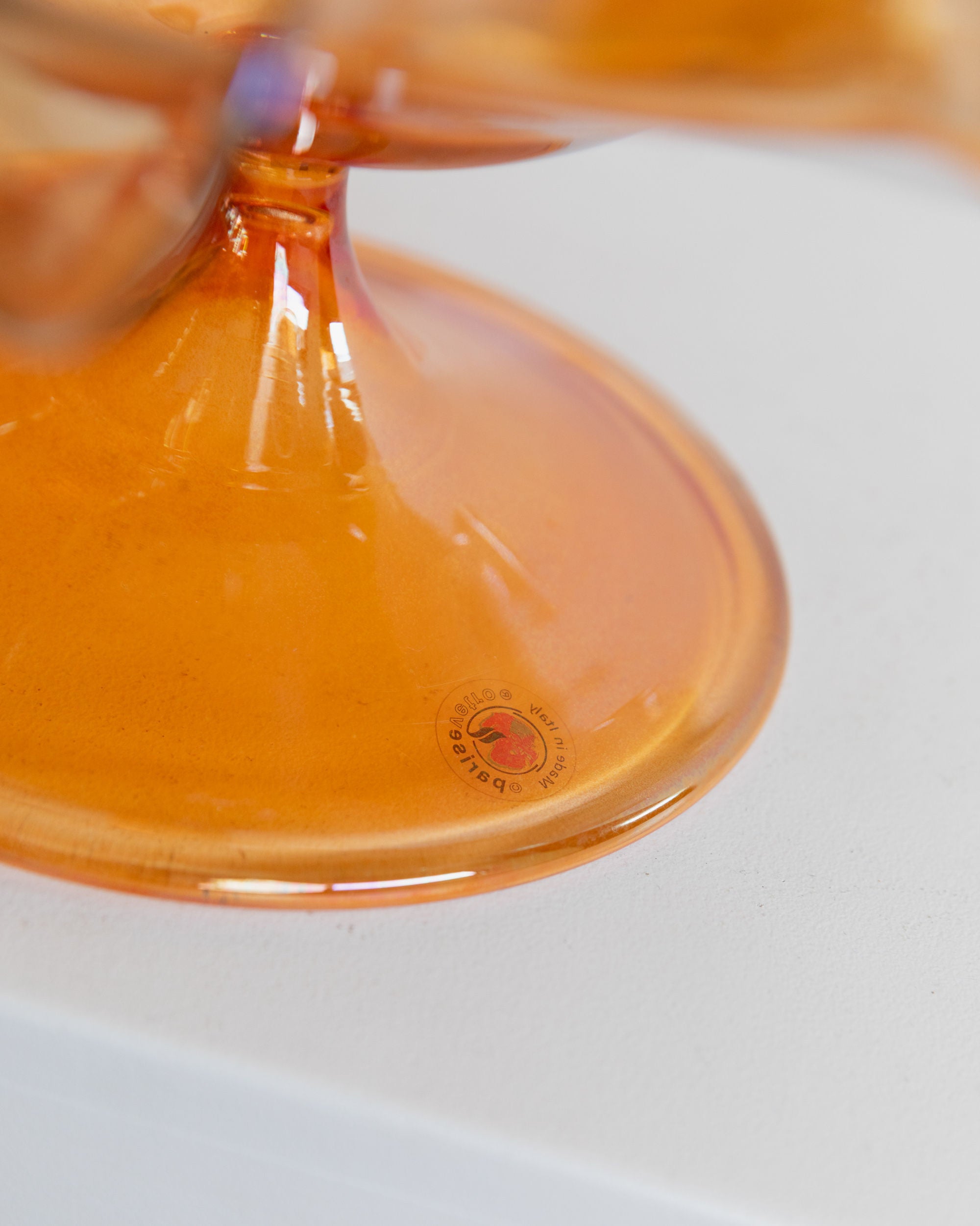 Close-up of a vintage Murano glass bowl by Parise Vetro from the 70s, featuring a rounded base with an orange iridescence. A small circular label displays "Bottega Jacobs." The background is a plain white surface, enhancing this exquisite decorative piece.