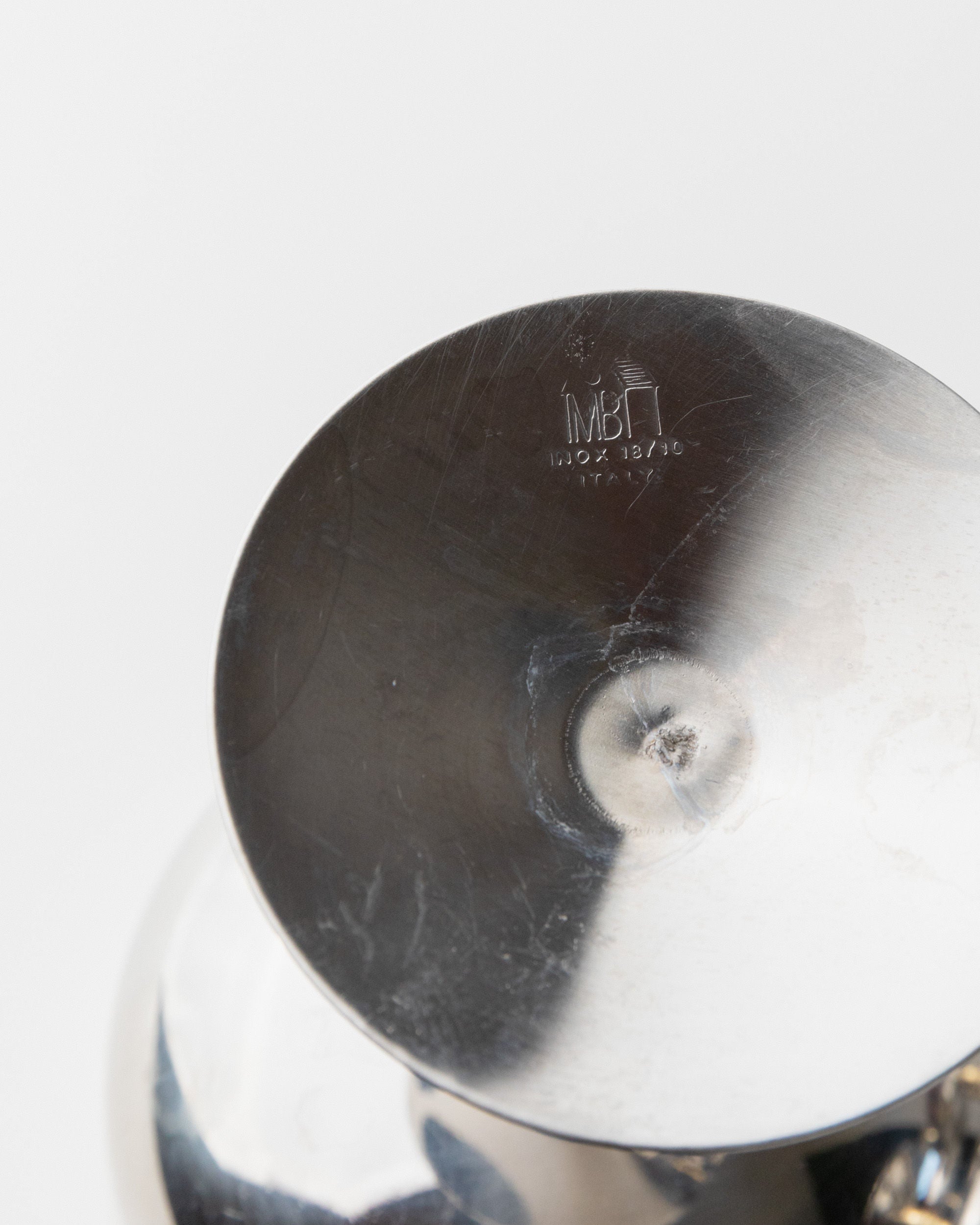 Close-up of the base of a metallic object displaying engraved markings: "IMBT," "100/2 18/76," and "Italy." The vintage surface of the Bottega Jacobs 6 Stainless Steel Champagne Coupes from the '70s is reflective with slight scratches.