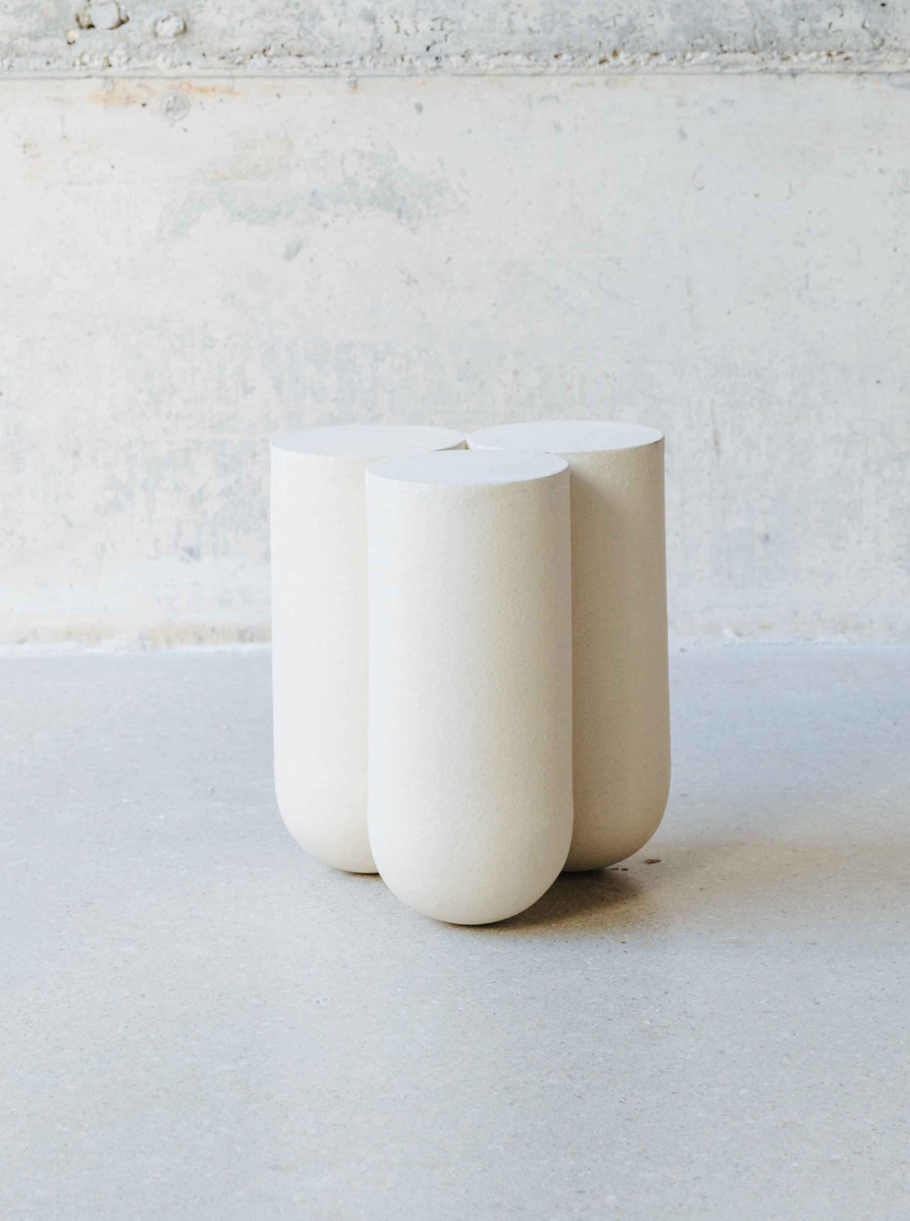 A minimalist, cream-colored Side Table S MOOR by Lisa Allegra, exhibiting a unique sculptural design with four rounded cylindrical legs that converge at the top. It is situated on a smooth, light-colored surface against a textured, partially painted wall and crafted from grogged stoneware.