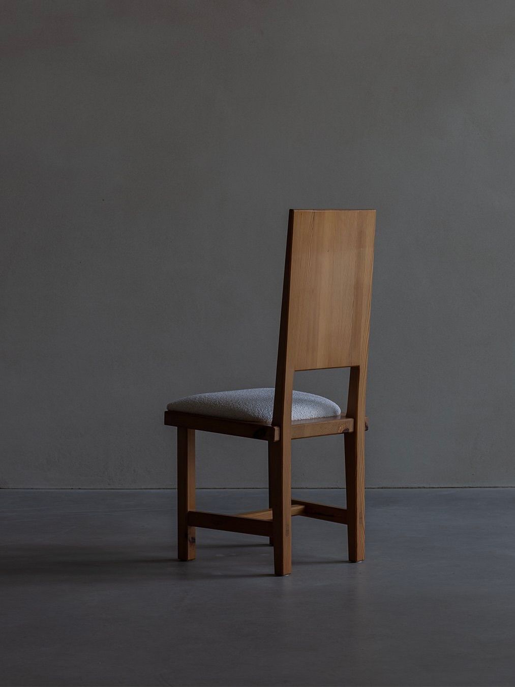 The Göran Malmvall - Model 553 chair from Bicci de' Medici features a wooden frame with a tall, straight backrest and a light gray cushioned seat. Set against a dark gray wall on a concrete floor, it embodies the simplicity and elegance of Swedish design.