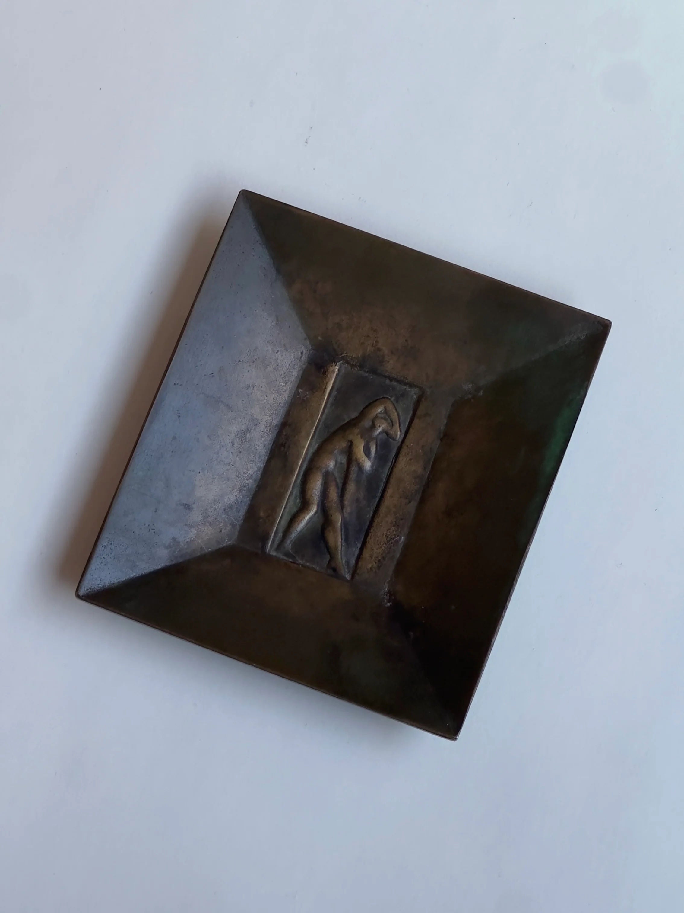 The Galerie Storm "Bronze dish, GAB" is a square sculpture with a recessed center showcasing a raised, stylized figure. Its dark, patina-coated surface is slightly reflective and minimalist, set against a plain light backdrop similar to Jacob Ängman's art style.