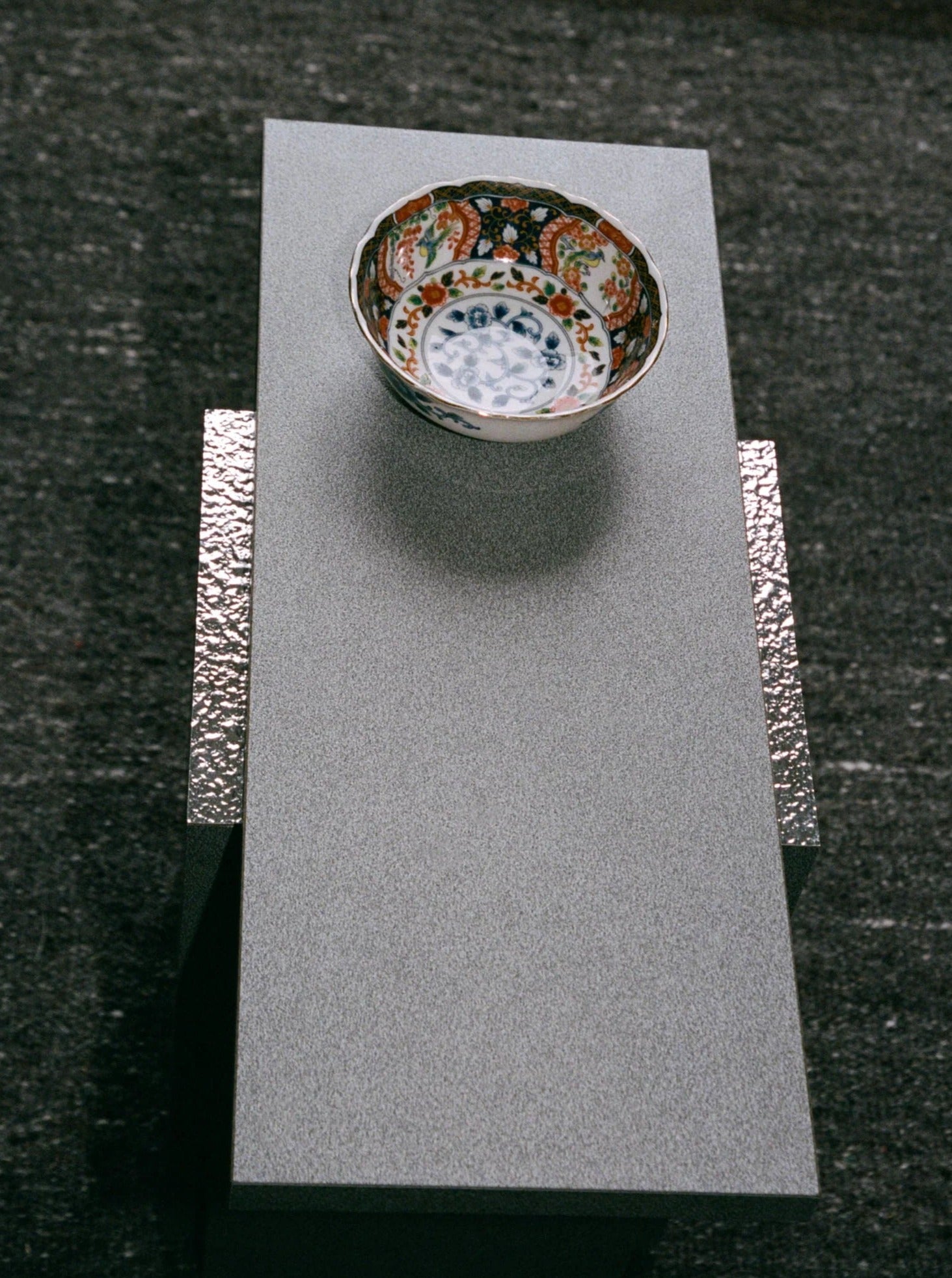 A decorative bowl with intricate patterns and vibrant colors is displayed on the "Mara T" Side Table – Silver Noise by TEGET, reminiscent of laminated furniture. The dark, textured background complements the subtle elegance of this setup.