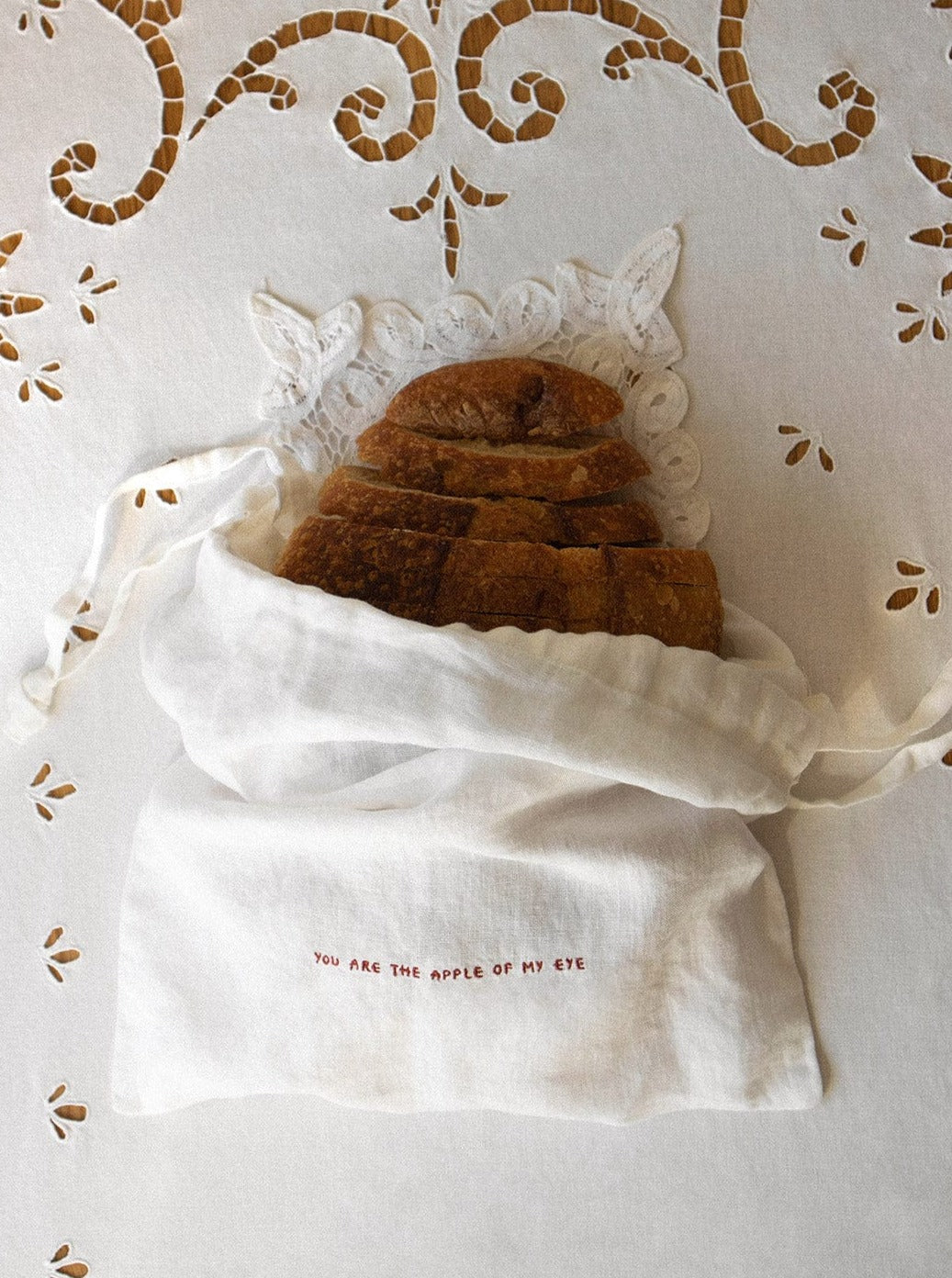 A crusty loaf of bread is artfully wrapped in the Reusable Linen Produce Bag 'You Are The Apple Of My Eye,' made by arkitaip. This OEKO-TEX certified linen bag keeps the bread fresh and features an embroidered message that reads "You are the apple of my eye." Both the bread and its elegant wrapping rest on a white linen tablecloth adorned with intricate cutwork and floral embroidery.