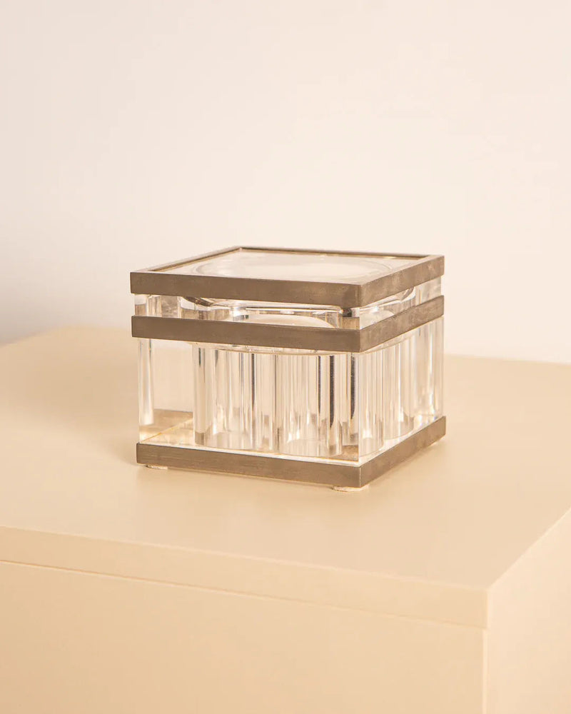 The Metal and Plexiglas box by Soldi & Scati 70's from Treaptyque, featuring a rectangular shape with metallic details and a Plexiglas body, rests on a light-colored surface, showcasing minimalist design. The box's clear sides and flat top highlight its elegance and simplicity.