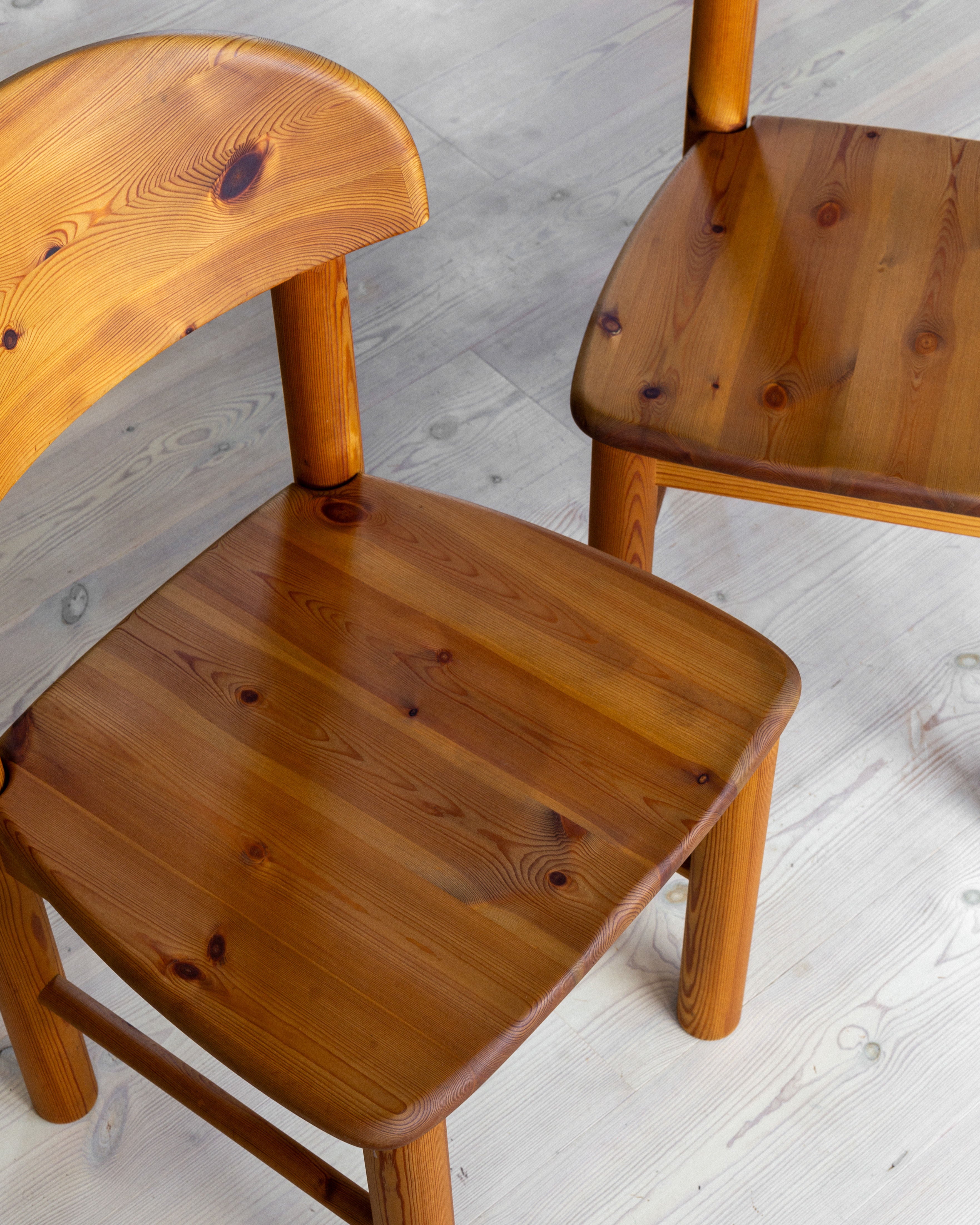 Two Spigel pine chairs by Rainer Daumiller, part of an iconic 1970s collection from Denmark, are positioned on a light-colored wooden floor. Crafted from solid pine, they feature curved backrests and smooth, polished seats that showcase a natural wood grain pattern, enhancing their rustic appearance. The composition highlights the close-up details of these Danish design masterpieces.