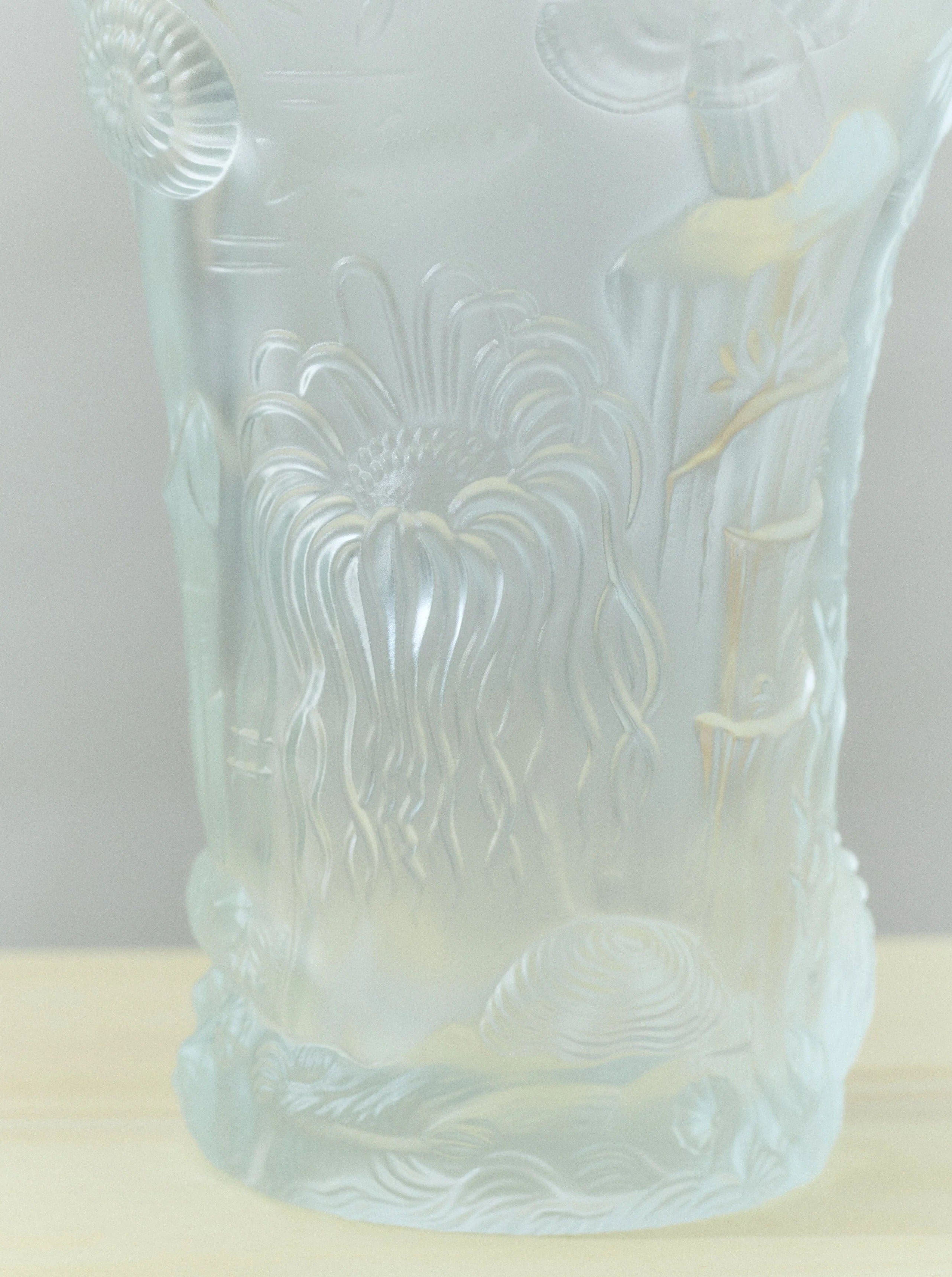 A detailed Michael Lagoon Vase from Akua Objects showcases intricate sea-themed carvings, including marine plants, shells, and a jellyfish. This handmade design wraps around the vase, creating a textured and elegant appearance. The Michael Lagoon Vase piece is placed on a plain, light-colored surface.