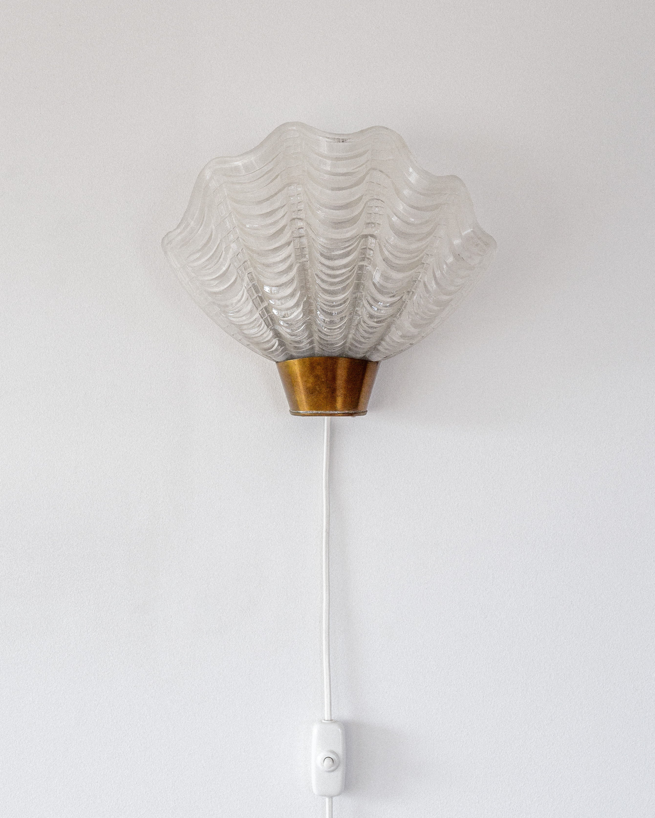 The Vintage "Coquille" Wall Light by Spigel, from Sweden 1940s, features a glass shell-like shade with a brass base mounted on a white wall. A stylish white cord with an attached switch enhances both the elegance and functionality of this fixture.