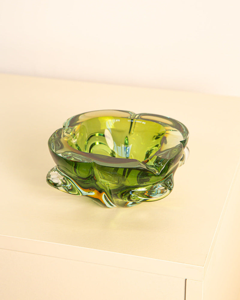The Treaptyque Dark Green Murano Glass “Fleur” Ashtray from the 60s, with an organic wavy form inspired by Italian design, is placed on a light beige surface against a pale background.
