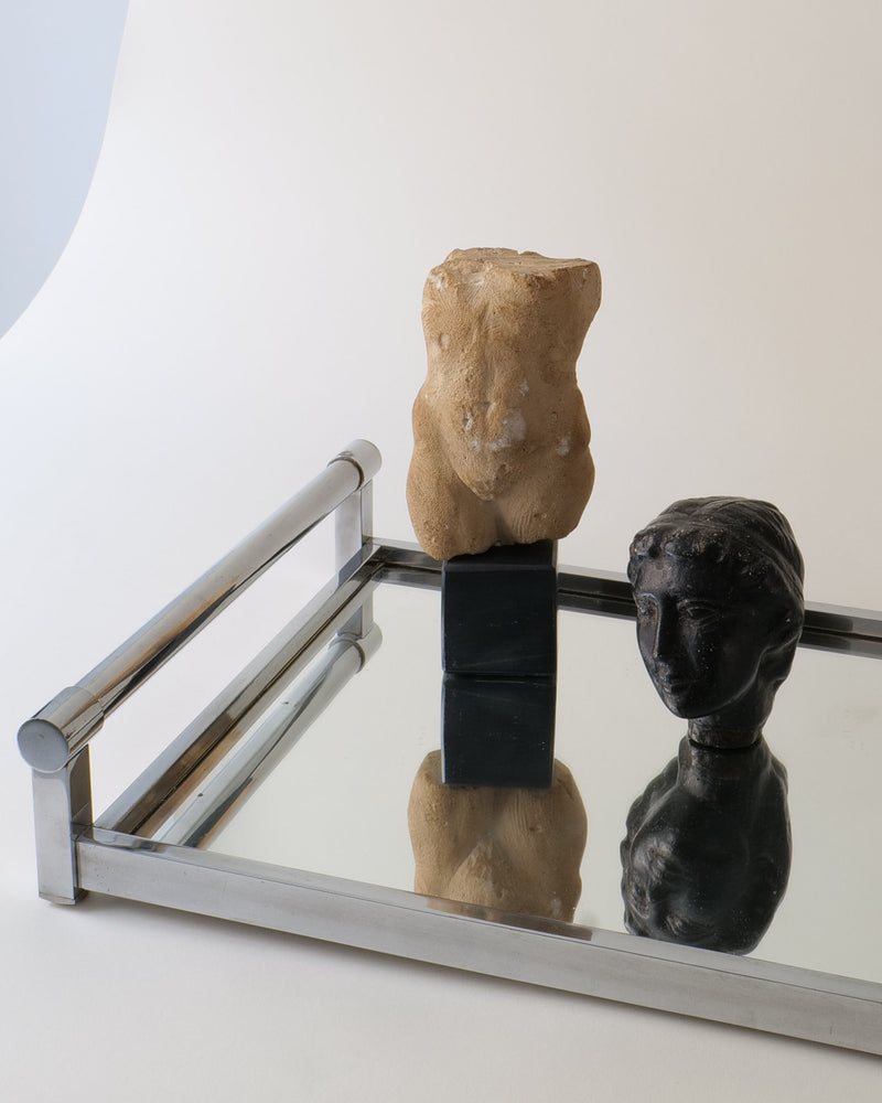 A mirrored tray displays the Torso Sculpture by Misja Kristoffer Rasmussen (Collection apart), beige and echoing Greek forms, beside a dark head bust—both reflected on the tray against a light background.