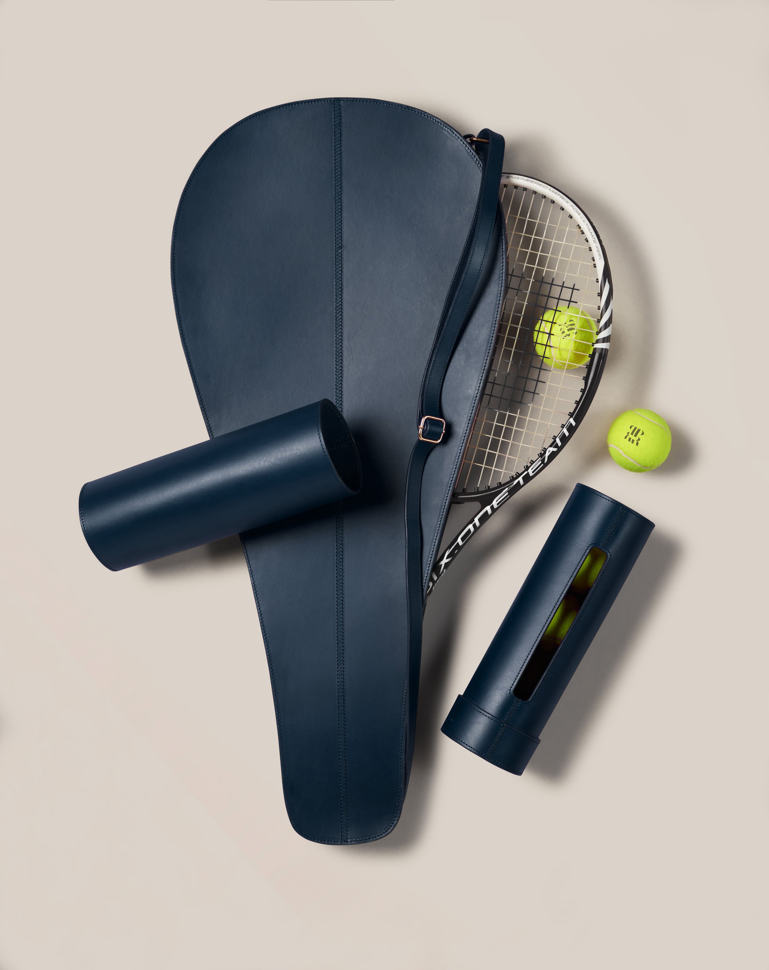 A Paradise Row Leather Tennis Racket Cover showcases a bespoke British design, partially enclosing a racket on a beige surface. Nearby, two tennis balls accompany one open and one closed cylindrical navy container.