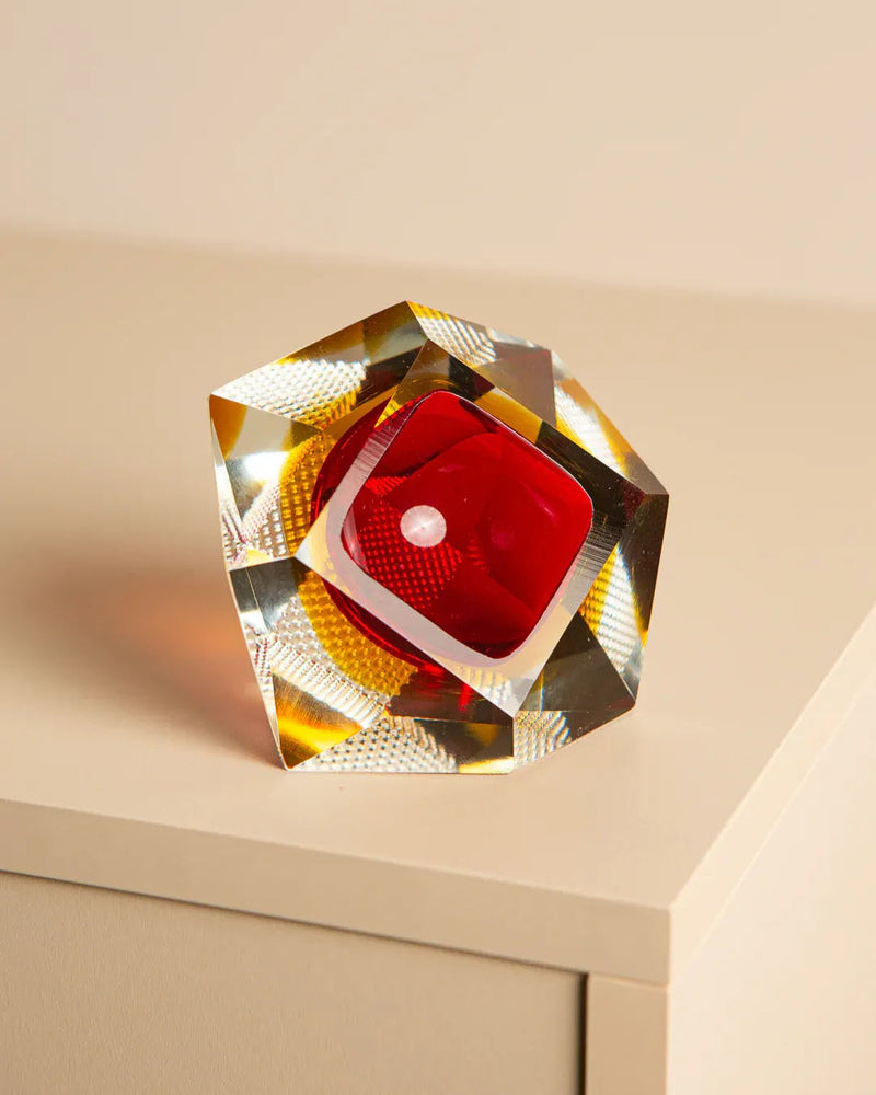 A colorful glass sculpture, similar to Flavio Poli's 'Diamant' Ashtray Murano for Seguso 60’s, rests on a white surface. It boasts a red core surrounded by clear and amber shades from Treaptyque, reflecting light elegantly in various directions.