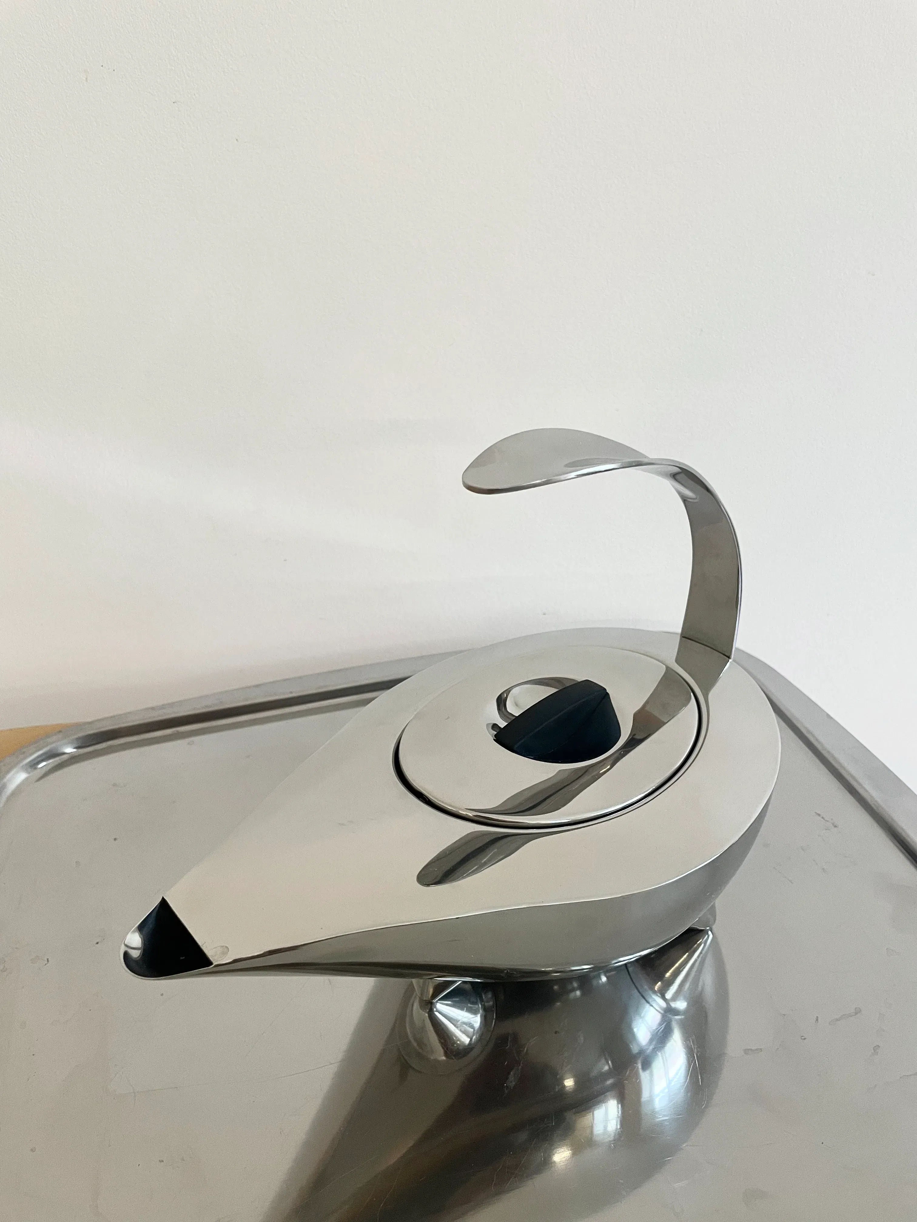 A sleek, modern Collect Cph 1980 Bodum Naoko stainless steel teapot with a curved handle and spout, sitting on a metal tray against a plain white wall.