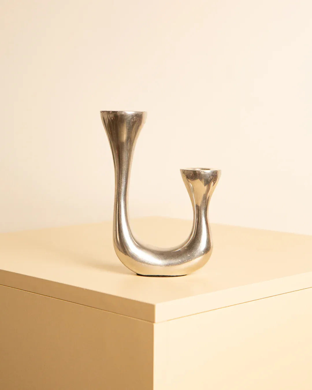 A sleek, avant-garde Treaptyque 80s Biomorphic Candleholders with two holders of varying heights forming a curvy U-shape, set on a light beige, minimalist tabletop against a matching beige background.