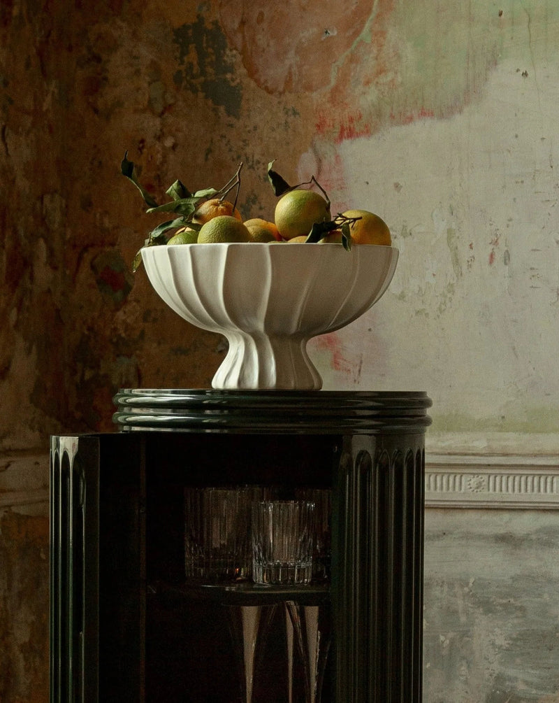 A Cloak Pedestal Bowl by Ransom & Dunn, part of a stunning ceramic collection handmade in Portugal, brimming with oranges and their leaves, sits atop a glossy black pedestal. The background features a rustic, textured wall with peeling paint in earthy tones.