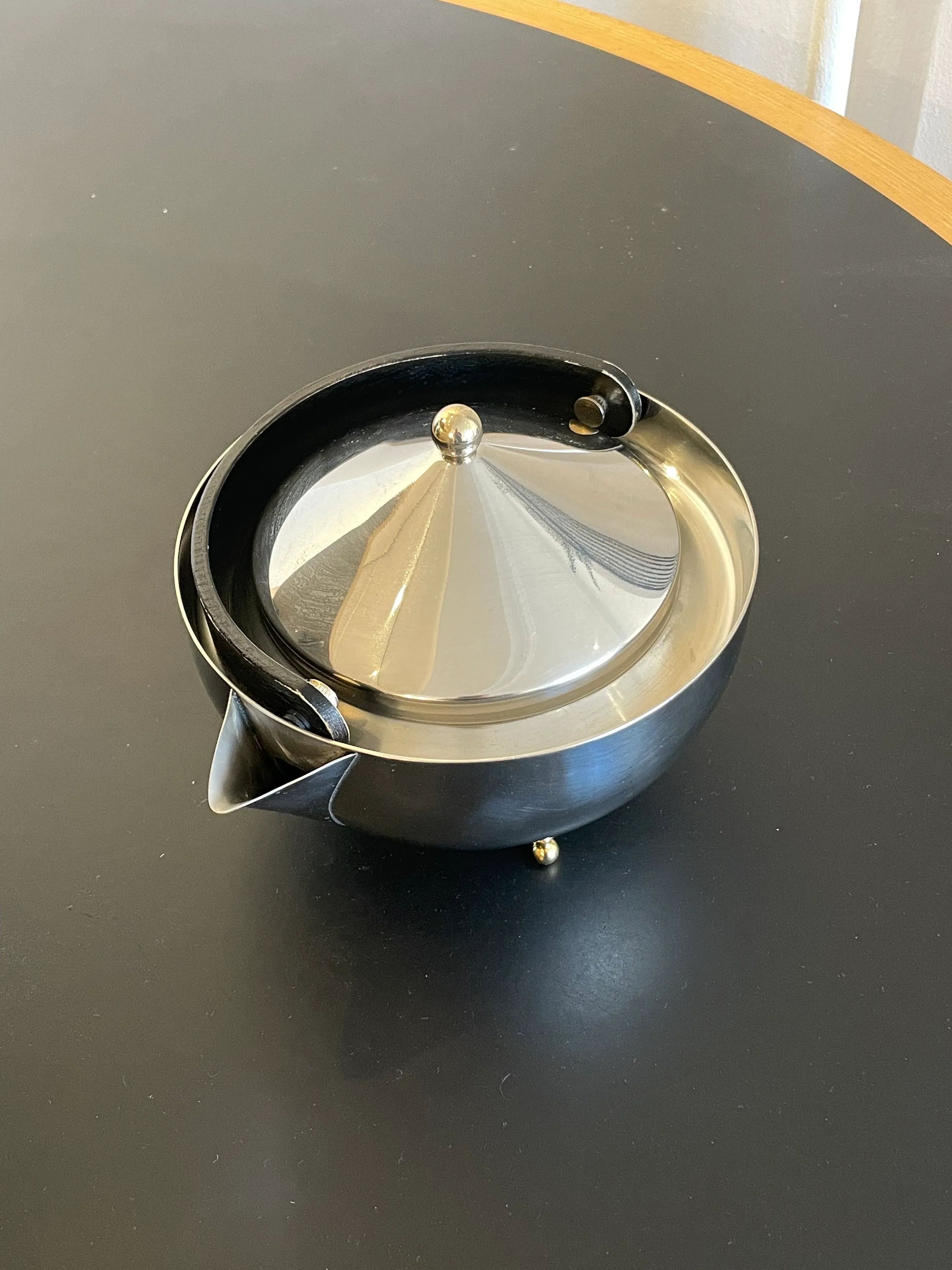 A sleek, modern Collect Cph Teaball Teapot with a conical lid and a black handle rests on a round, dark surface. The stainless steel teapot features a small spout and golden knobs on the lid and base. The setting appears minimalistic with a hint of a wooden edge in the background.