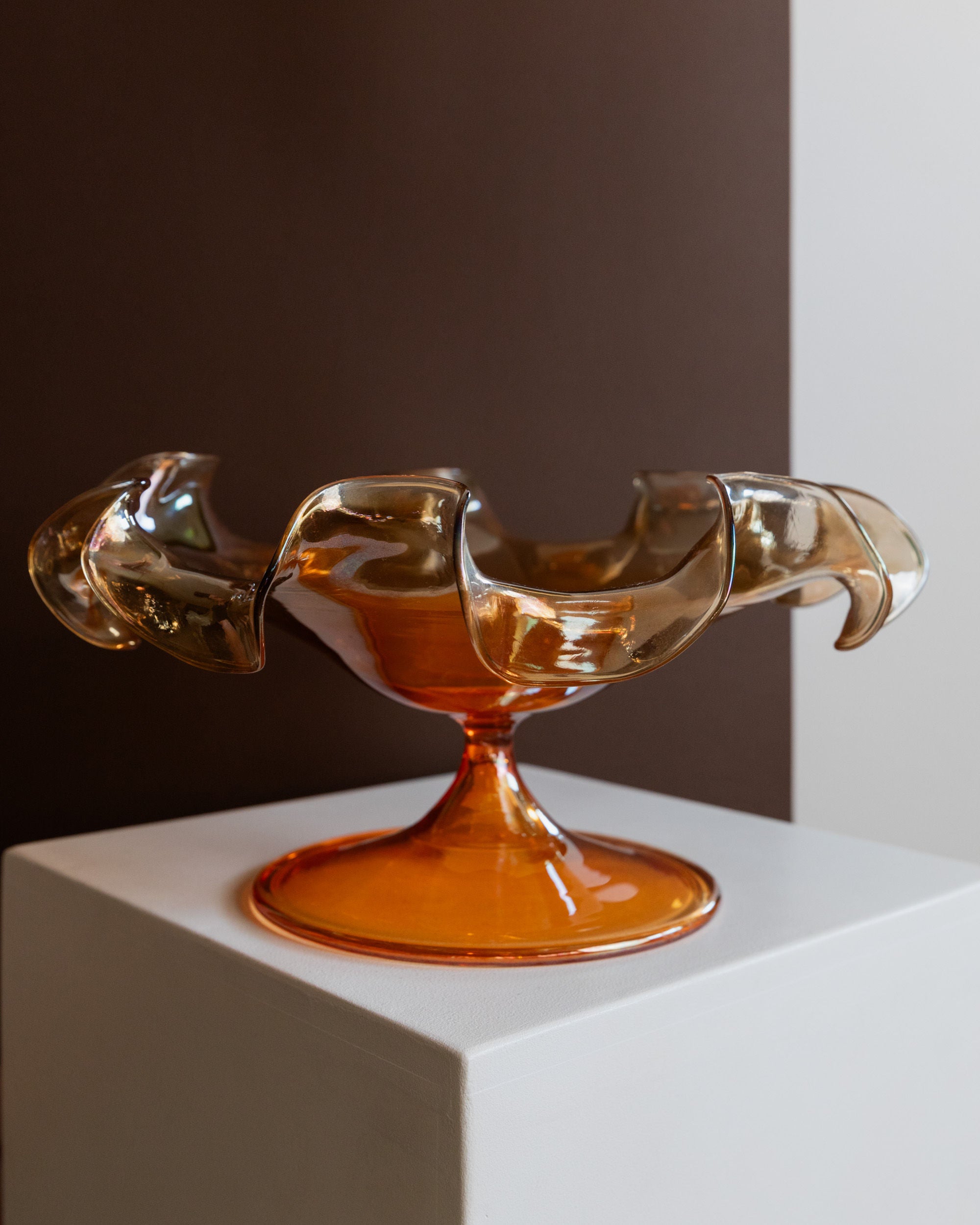 A translucent, orange Murano Glass Bowl by Parise Vetro 70s from Bottega Jacobs, featuring wavy, ruffled edges, sits on a white pedestal. The background boasts a split design with dark brown and beige tones that accentuate the artistic curves and vintage charm of this decorative piece.