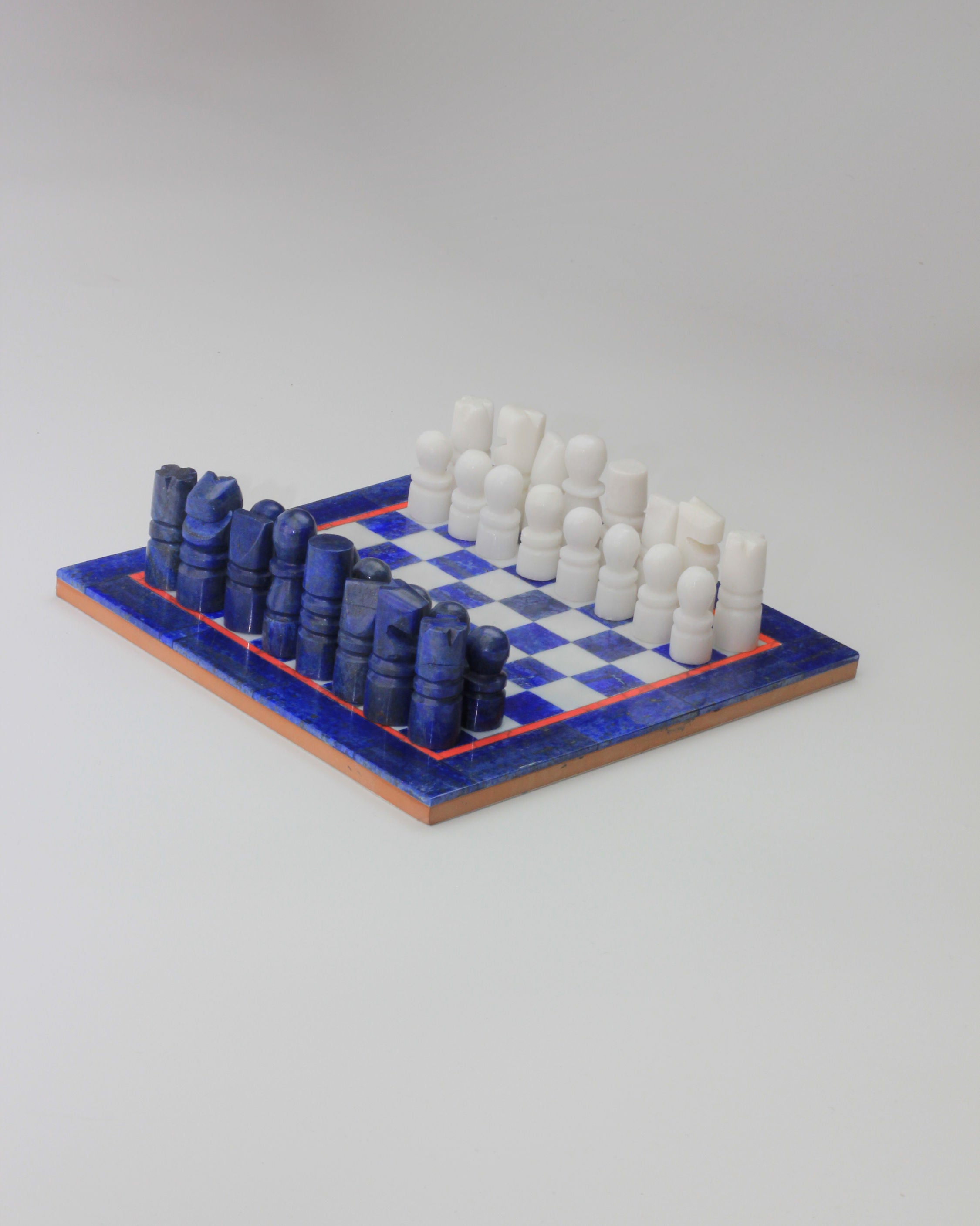 The Lapis Lazuli Chessboard from Boga Avante Shop features blue and white pieces arranged on a matching checkered board with a thin orange border. The minimalist design of this vintage chess set is placed on a light grey background, offering an elegant and timeless appeal.