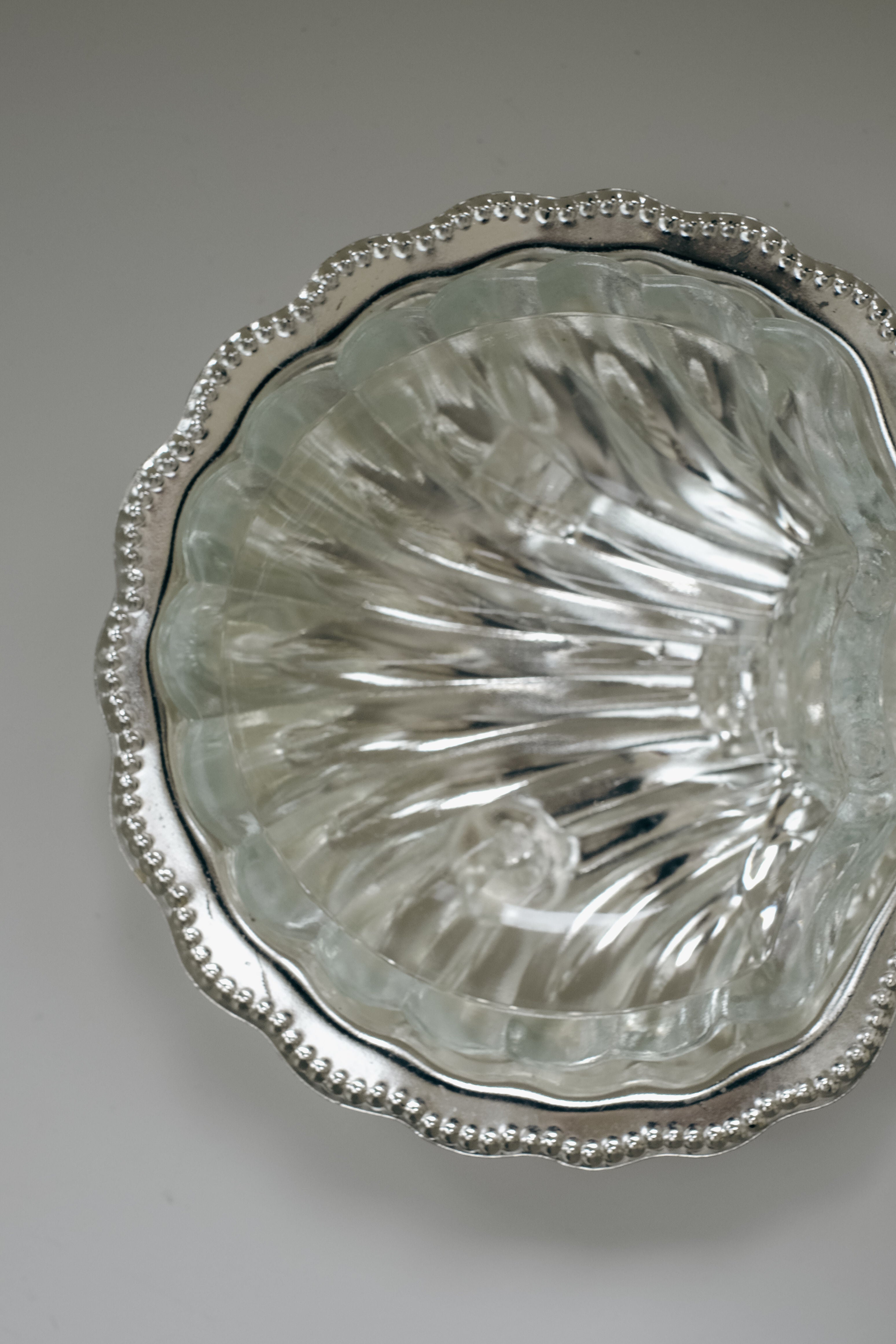 A Shell Shaped Butter Dish by septembre studios, with a reflective beaded silver rim and wavy scalloped edge, sits on a light gray surface. Its clear glass is patterned with vertical ridges.